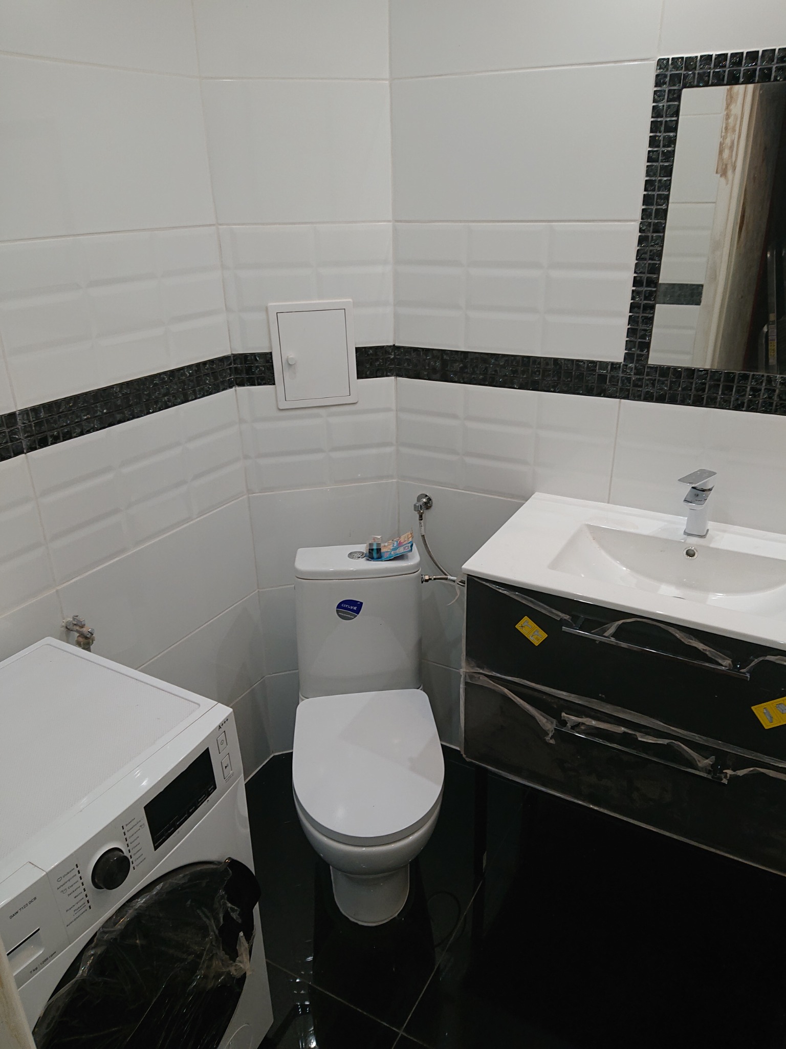 Biała łazienka z toaletą marki Cersanit, umywalką z czarną szafką w folii ochronnej i pralką, wykończona białymi płytkami z czarnym pasem mozaiki.