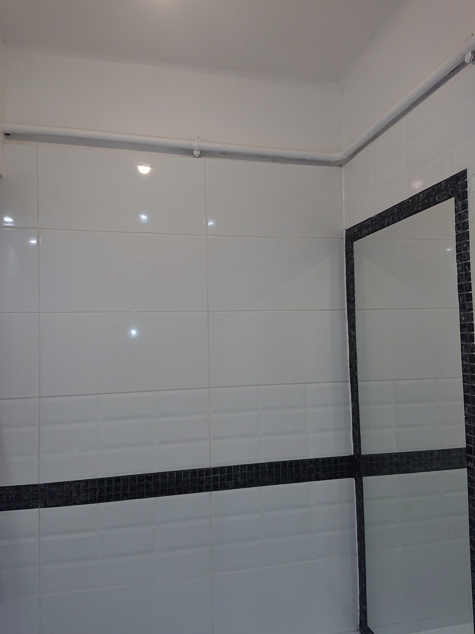 Białe, prostokątne płytki z czarnym pasem mozaiki w wyremontowanej łazience, widoczna rura pod sufitem i fragment lustra z czarną ramą.
