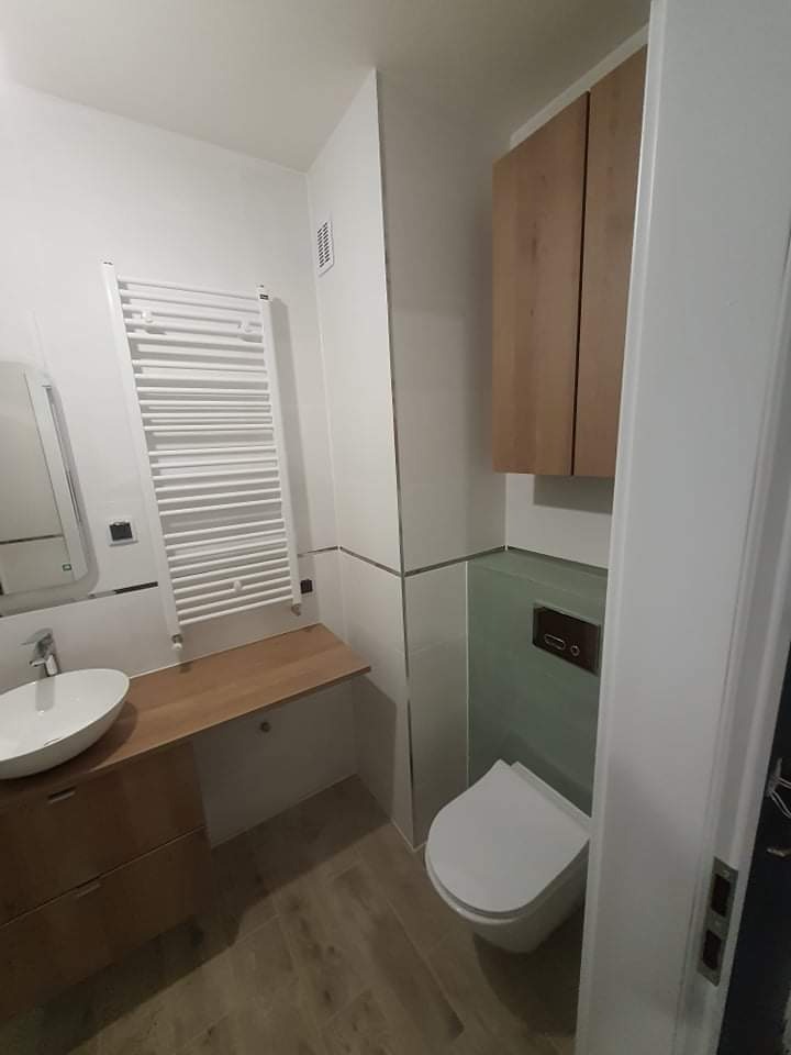 Nowoczesna łazienka z drewnianymi akcentami: szafka pod umywalką z blatem, wisząca toaleta z zielonymi płytkami, grzejnik drabinkowy i drewniana szafka wisząca.