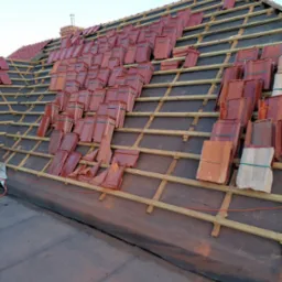 Częściowo pokryty dach z czerwonej dachówki ceramicznej, widoczne łaty i kontrłaty, przygotowane pakiety dachówek na dachu, komin w tle.