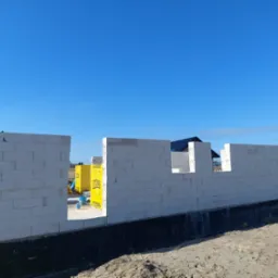 Wznoszone ściany domu z bloczków Ytong na fundamencie z widocznym hydroizolacją, w tle błękitne niebo i zabudowa jednorodzinna.