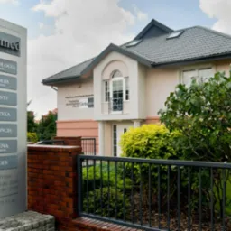 Budynek kliniki z szyldem informacyjnym o usługach: stomatologia, chirurgia, implanty, protetyka, ortodoncja, higiena, rentgen. Widoczne godziny przyjęć i numer telefonu. Ogród z krzewami...