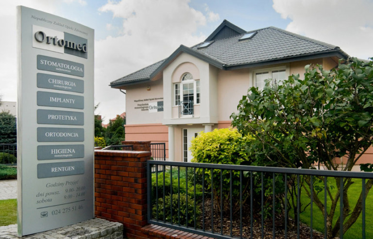 Budynek kliniki z szyldem informacyjnym o usługach: stomatologia, chirurgia, implanty, protetyka, ortodoncja, higiena, rentgen. Widoczne godziny przyjęć i numer telefonu. Ogród z krzewami...