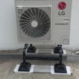 Zewnętrzna jednostka pompy ciepła LG Therma V zamontowana na czarnych wspornikach, z taśmą zabezpieczającą na betonowych płytach, na tle białej ściany. Widoczny fragment chodnika i ziemi.