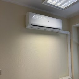 Global Green Energy Sp. z o.o. - Biały klimatyzator Haier zamontowany na beżowej ścianie w pomieszczeniu z oświetleniem sufitowym i widoczną instalacją odprowadzającą skropliny w białej rurze PCV.