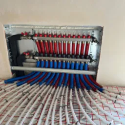 Rozdzielacz ogrzewania podłogowego z rurami PEX ułożonymi na folii aluminiowej z siatką, widoczne zawory regulacyjne i odpowietrzniki.