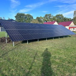 Global Green Energy Sp. z o.o. - Rząd paneli słonecznych na metalowych podporach, świeżo zainstalowany na trawniku, z widocznymi śladami ziemi i cieniem osoby w prawym dolnym rogu, domy w tle pod błękitnym niebem.