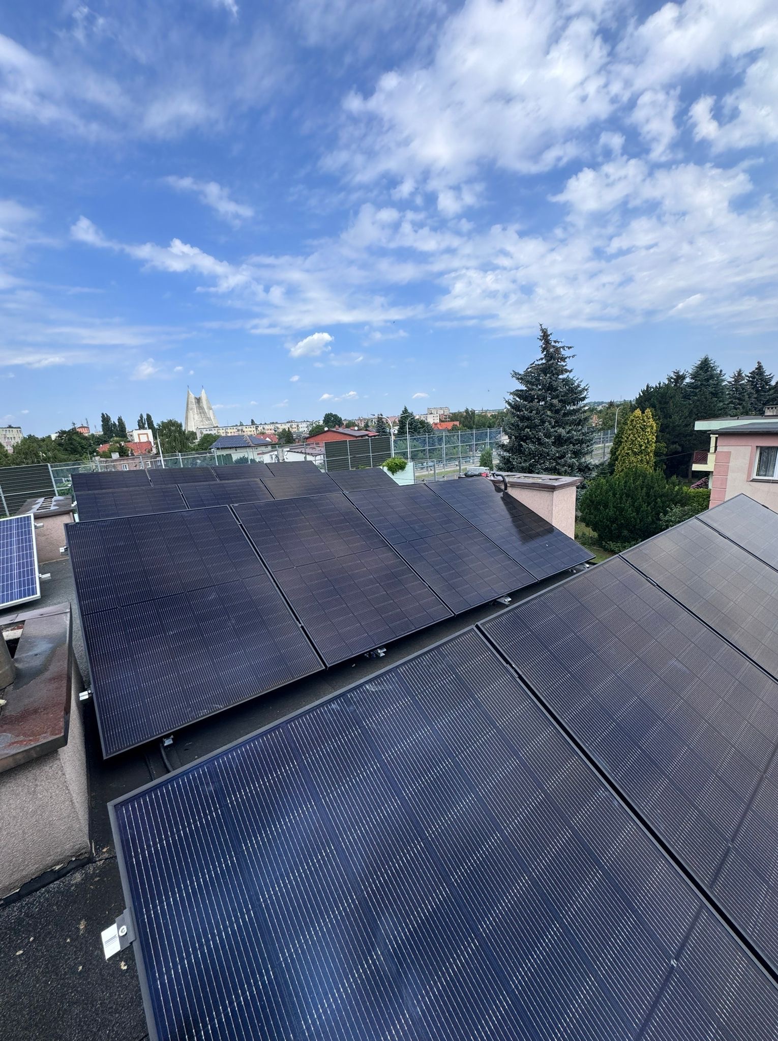Panele słoneczne na dachu budynku z widokiem na miasto i błękitne niebo z chmurami.