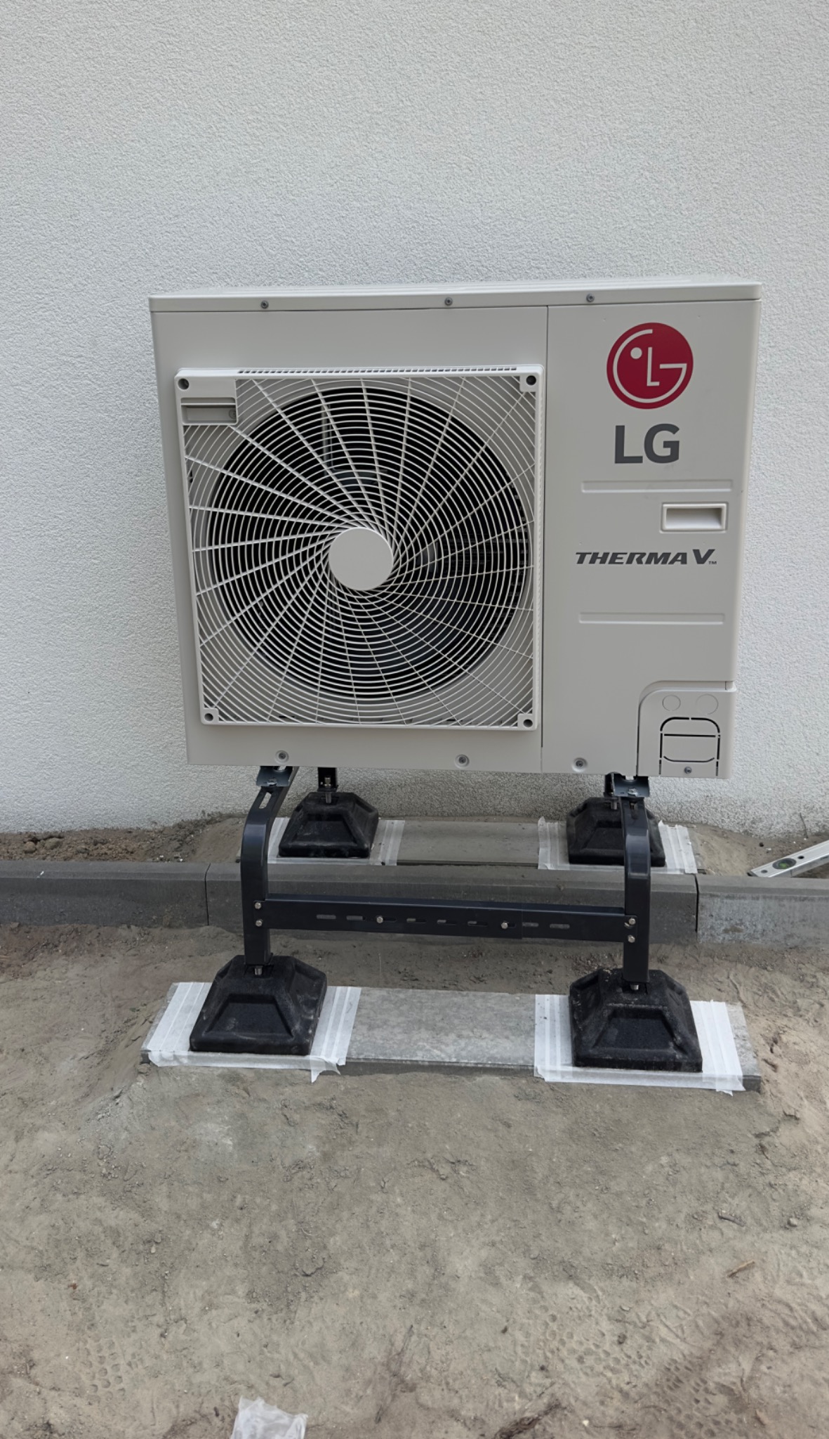 Zewnętrzna jednostka pompy ciepła LG Therma V zamontowana na czarnych wspornikach, z taśmą zabezpieczającą na betonowych płytach, na tle białej ściany. Widoczny fragment chodnika i ziemi.