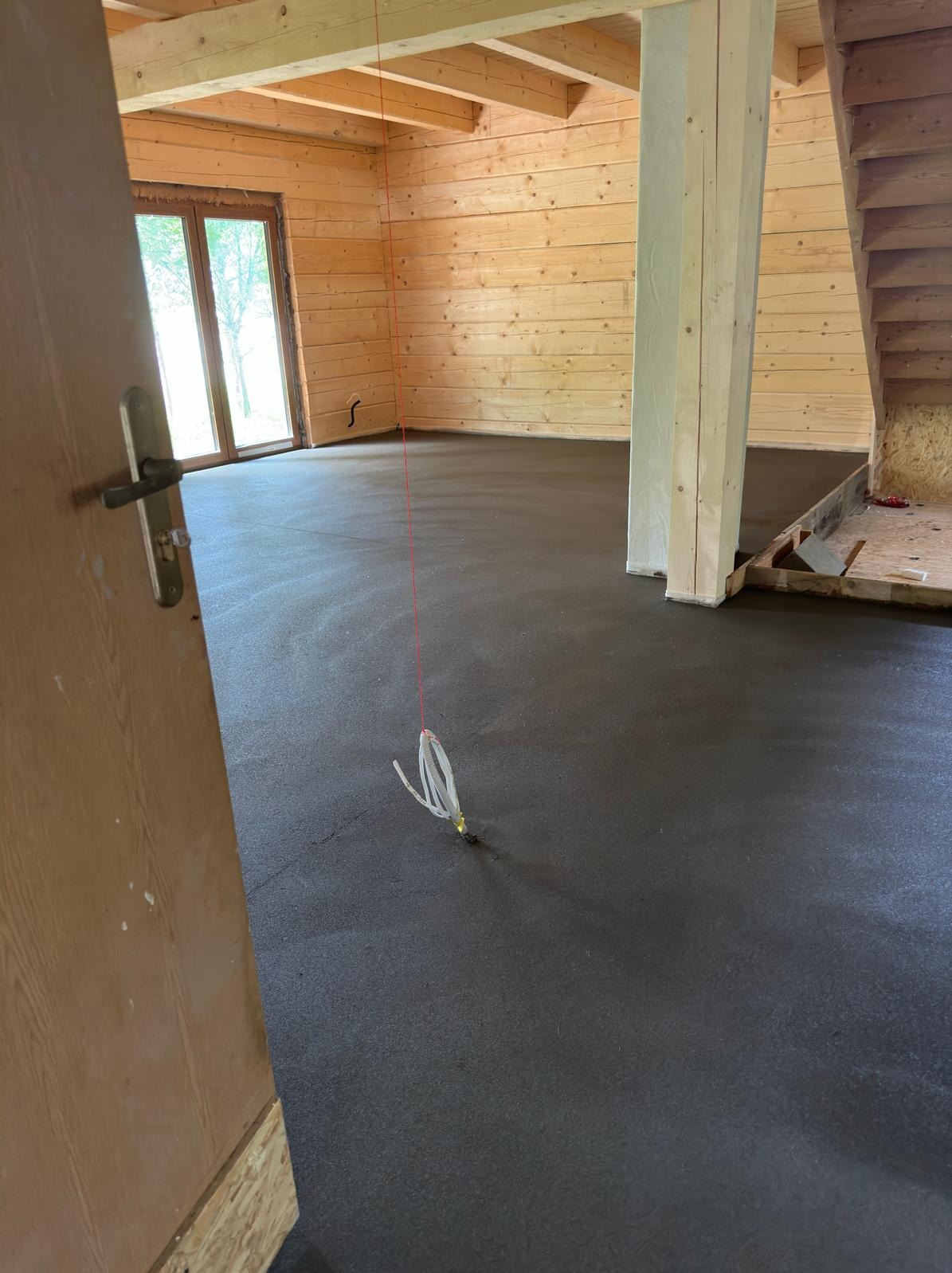 Wnętrze drewnianego domu w trakcie wylewki pod ogrzewanie podłogowe, widoczne rurki systemu grzewczego, surowy stan wykończenia, drewniane ściany i schody.