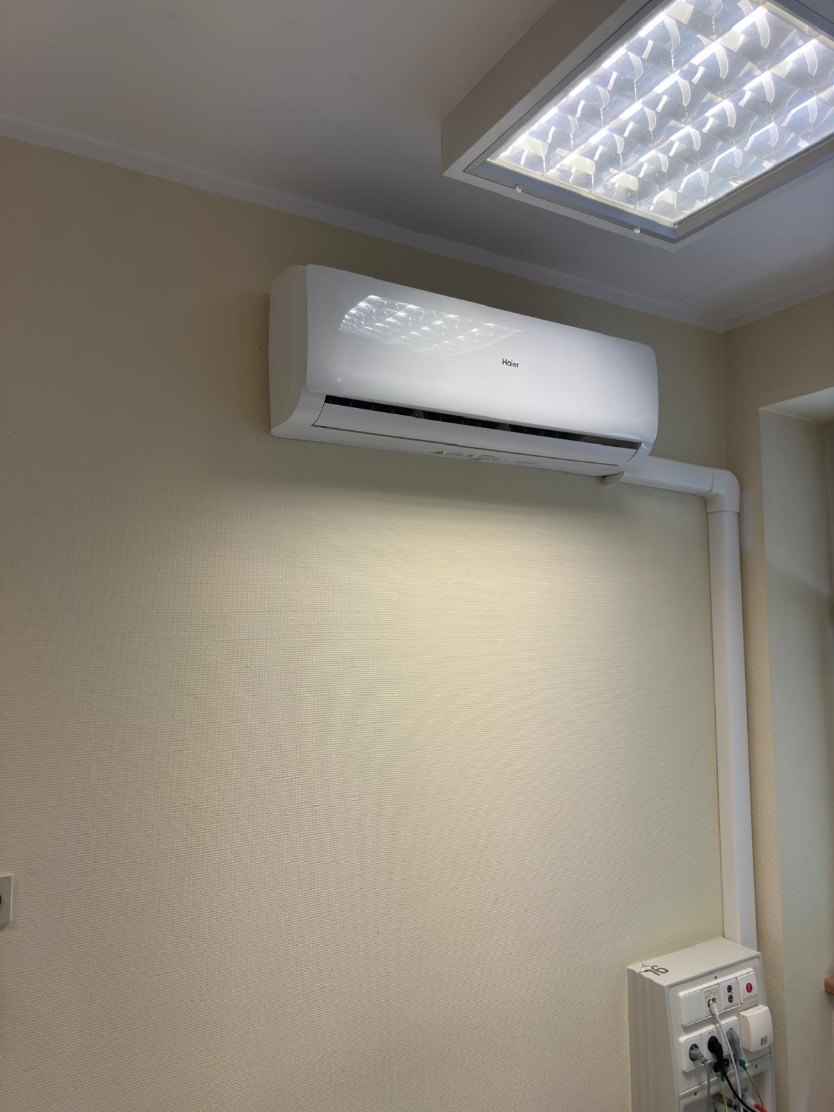 Biały klimatyzator Haier zamontowany na beżowej ścianie w pomieszczeniu z oświetleniem sufitowym i widoczną instalacją odprowadzającą skropliny w białej rurze PCV.