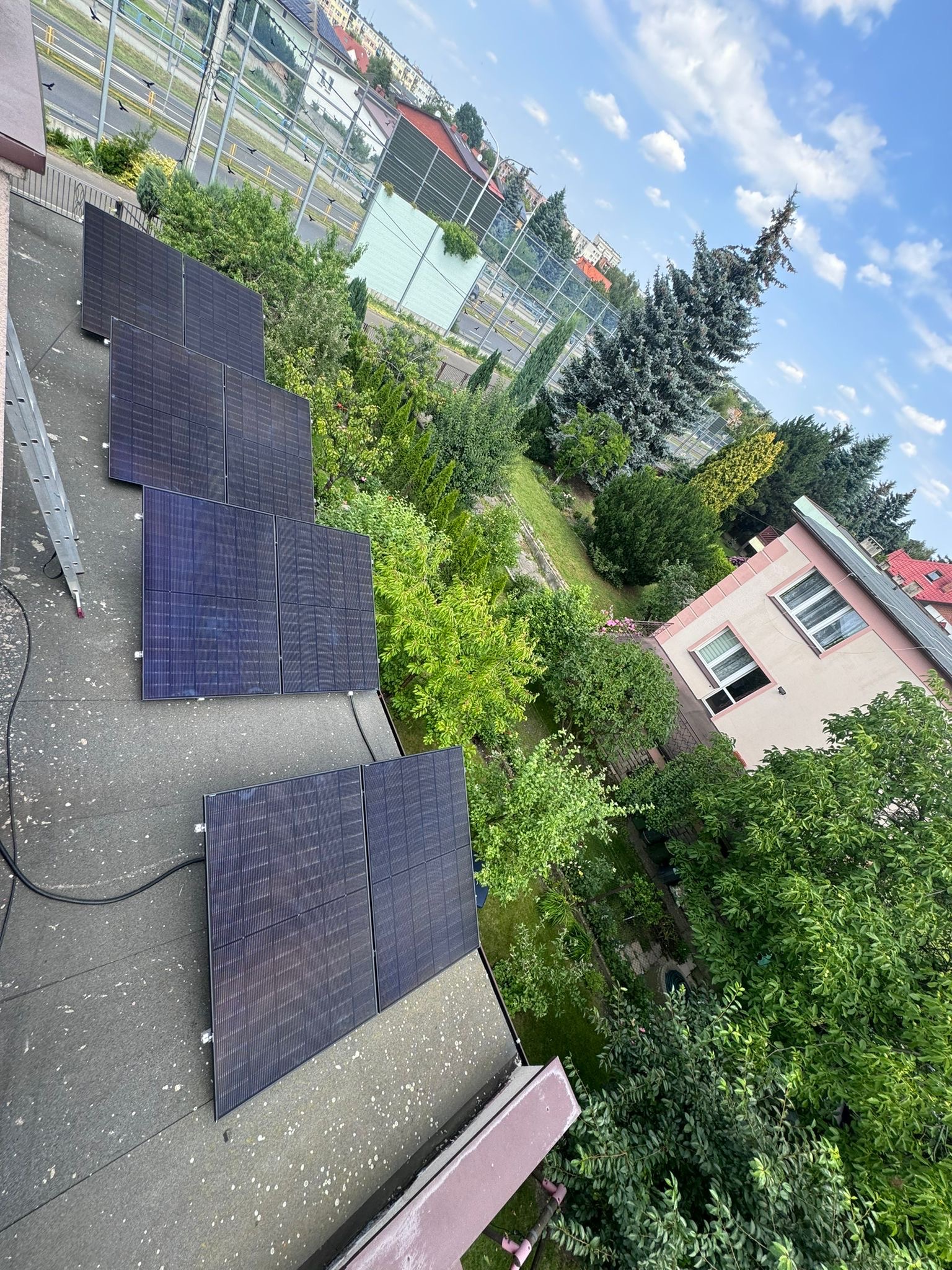 Instalacja paneli słonecznych na płaskim dachu budynku, widoczna drabina, w tle zieleń drzew i zabudowa miejska pod błękitnym niebem.