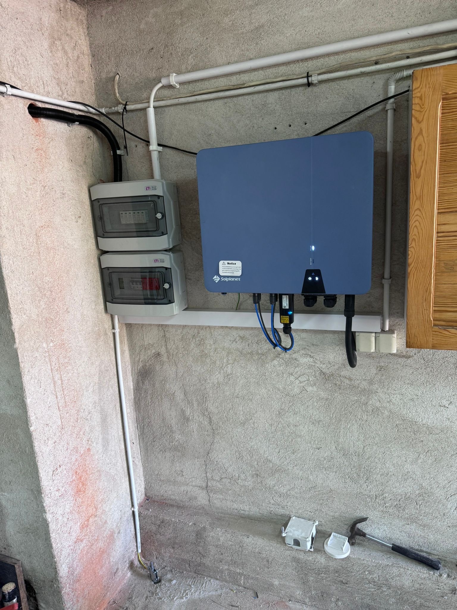 Niebieski inwerter fotowoltaiczny marki Solplanet zamontowany na ścianie z widocznymi przewodami, obok skrzynki bezpiecznikowe ETI i fragment szafki z drewna. Na podłodze leżą elementy instalacyjne...
