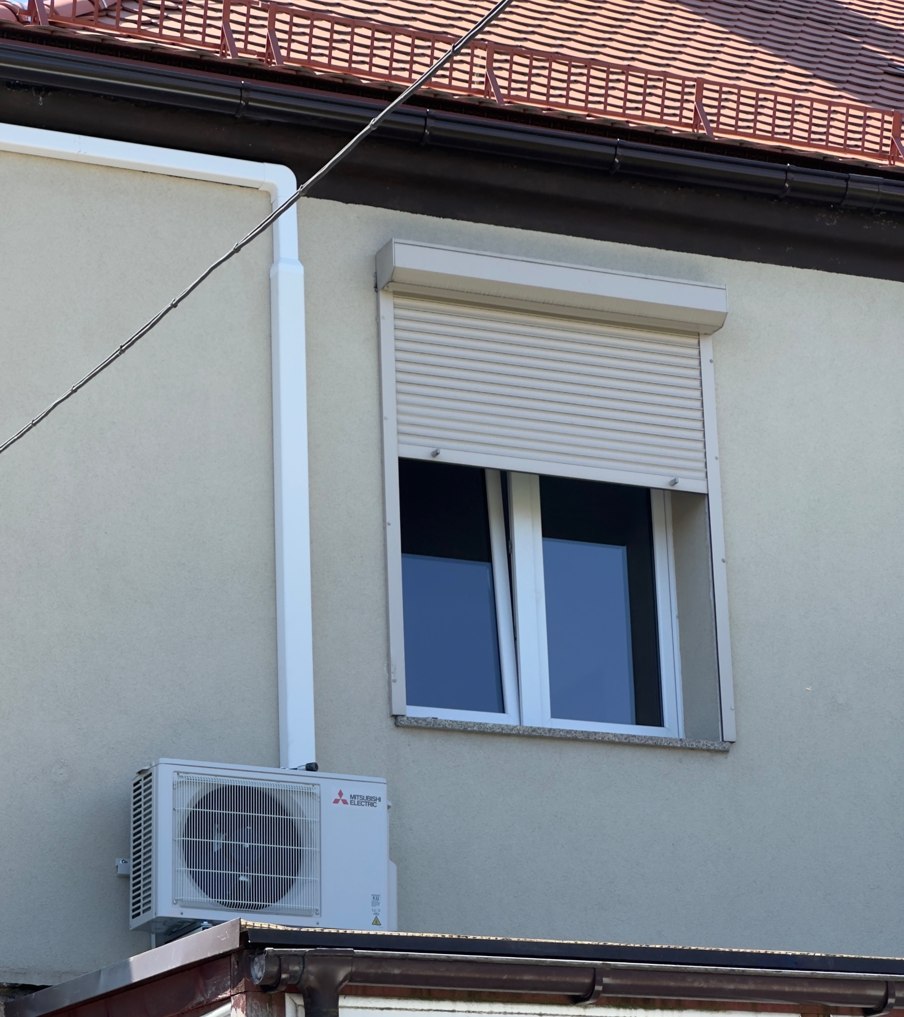 Zewnętrzna jednostka klimatyzacji Mitsubishi Electric zamontowana na elewacji budynku, obok okna z opuszczoną roletą.