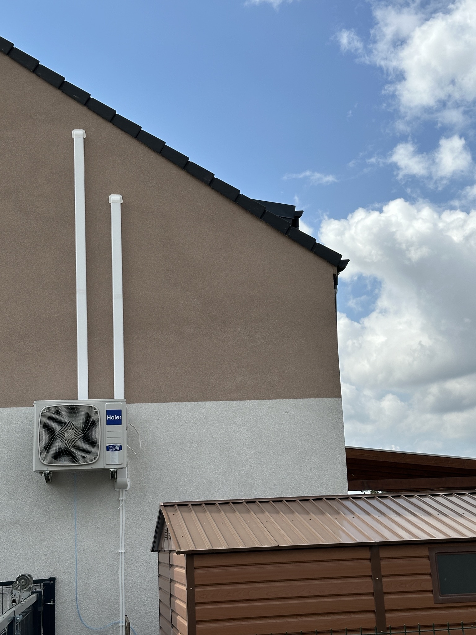 Zewnętrzna jednostka klimatyzatora Haier zamontowana na dwukolorowej elewacji domu z widocznymi rurami odprowadzającymi, na tle błękitnego nieba z cumulusami i brązowej blaszanej budki.