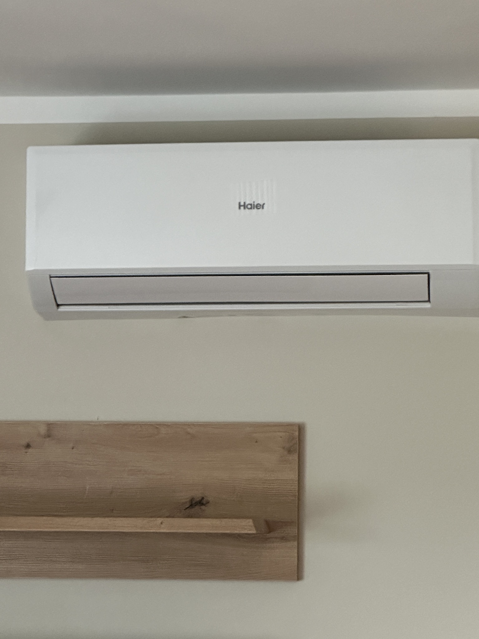 Biały klimatyzator Haier zamontowany na ścianie nad drewnianą półką. Widoczny z bliska, w neutralnym oświetleniu.