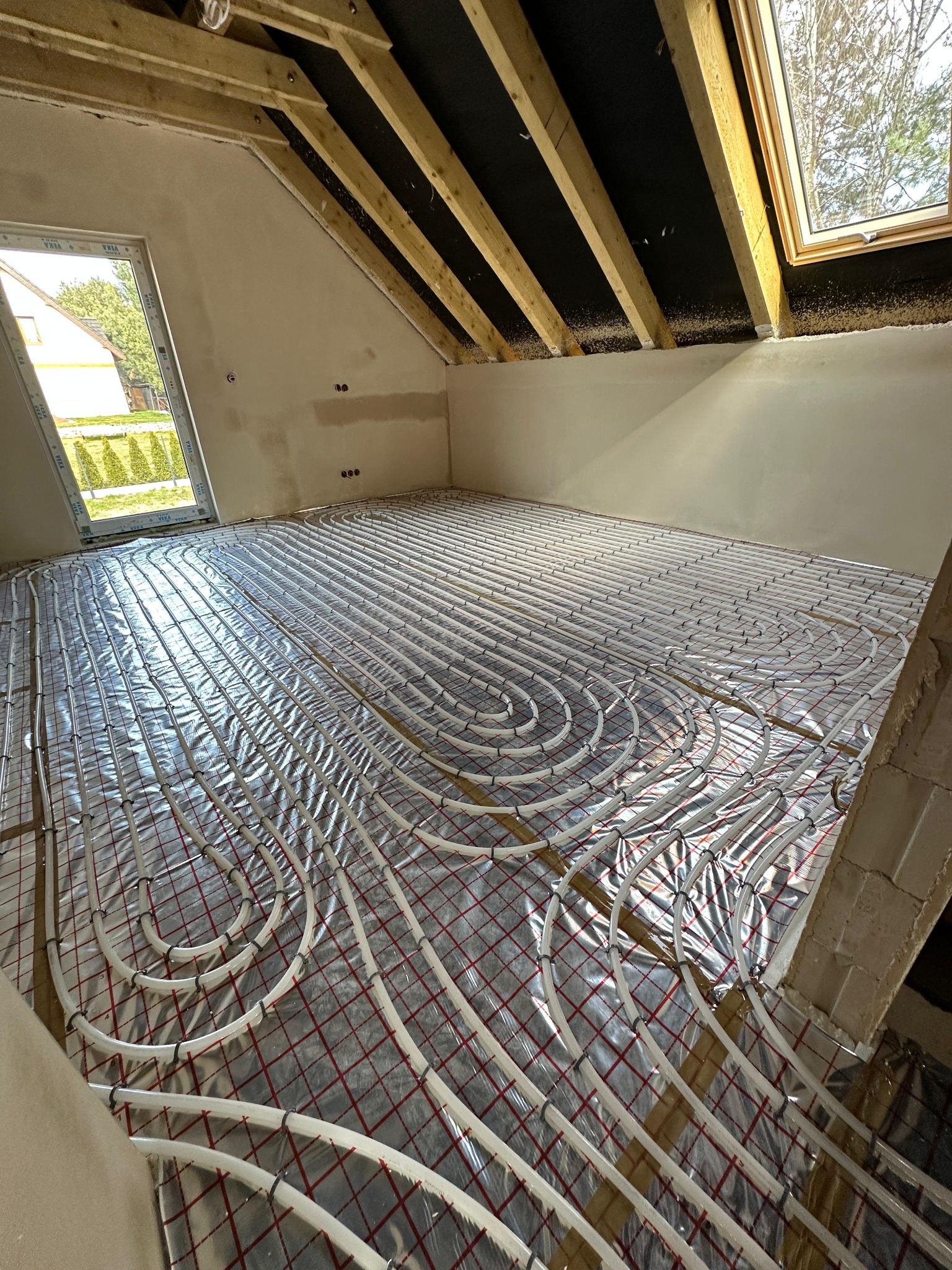 Instalacja ogrzewania podłogowego w poddaszu, widoczne białe rury ułożone na folii z czerwoną siatką, okna dachowe w tle, surowy stan ścian.