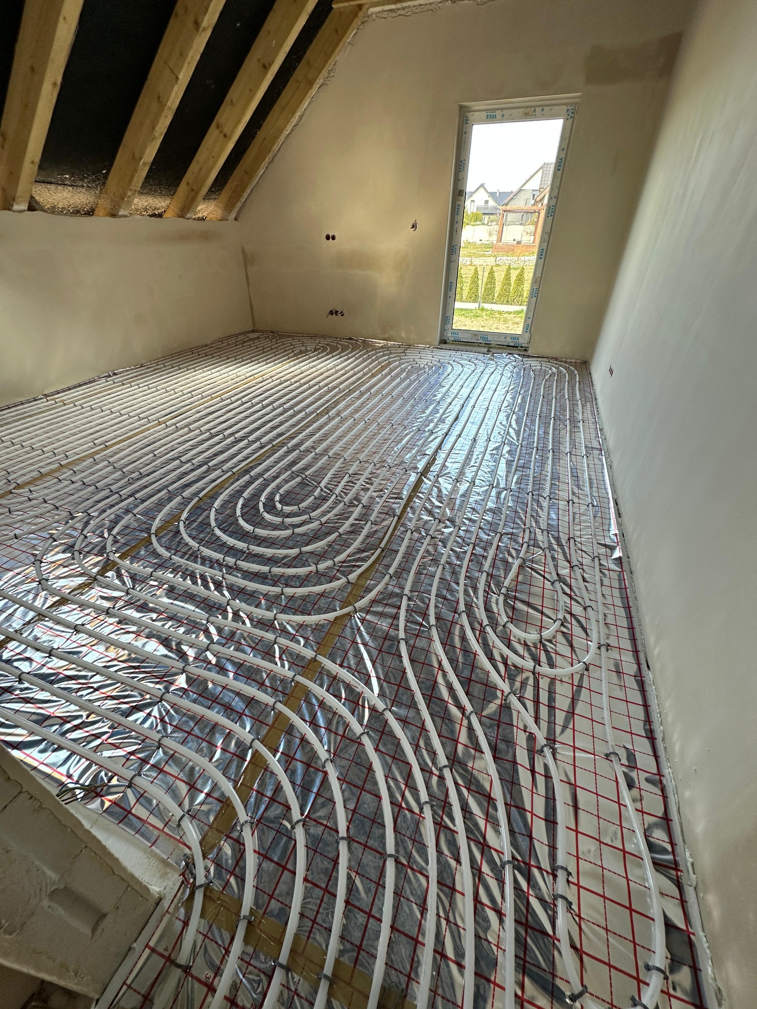 Białe rury ogrzewania podłogowego ułożone na srebrnej folii z czerwoną siatką w surowym pomieszczeniu z oknem i drewnianym stropem.