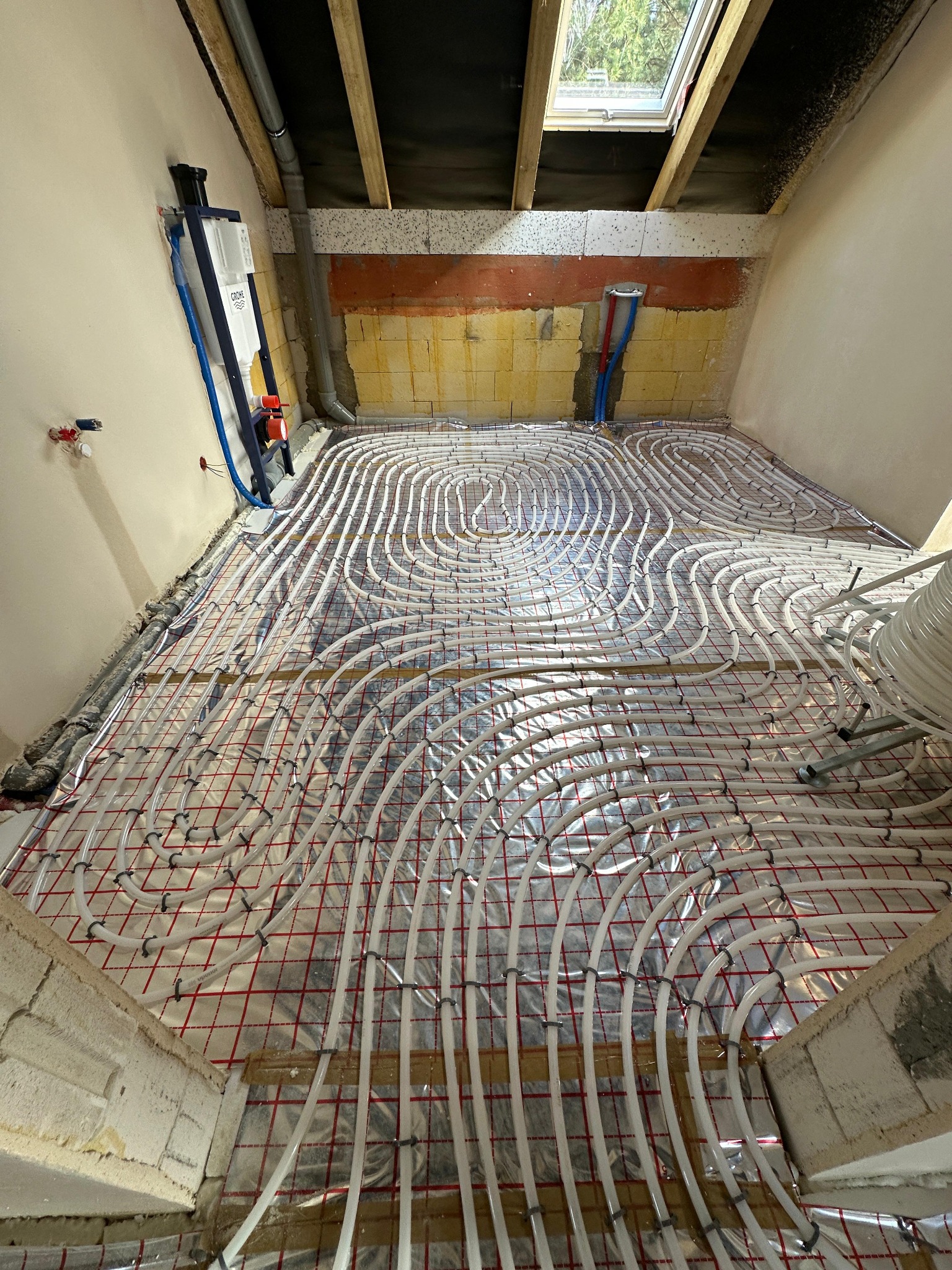 Układanie białych rur grzewczych na folii odblaskowej z czerwoną siatką na podłodze w trakcie montażu ogrzewania podłogowego w pomieszczeniu z oknem dachowym i instalacją sanitarną na ścianie.