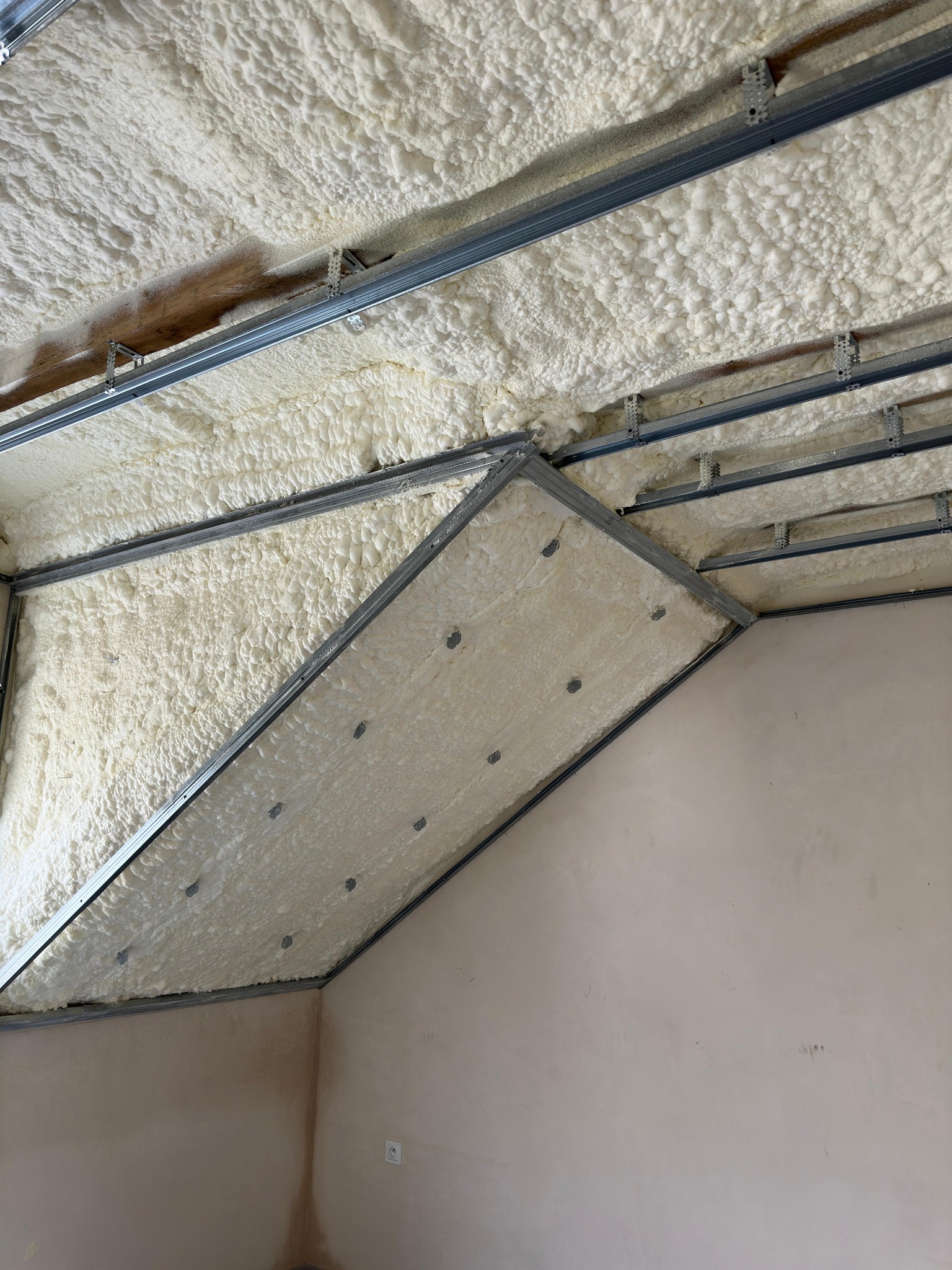 Poddasze z widoczną pianą izolacyjną na suficie i częściowo na ścianie szczytowej, zamontowaną na metalowym stelażu.