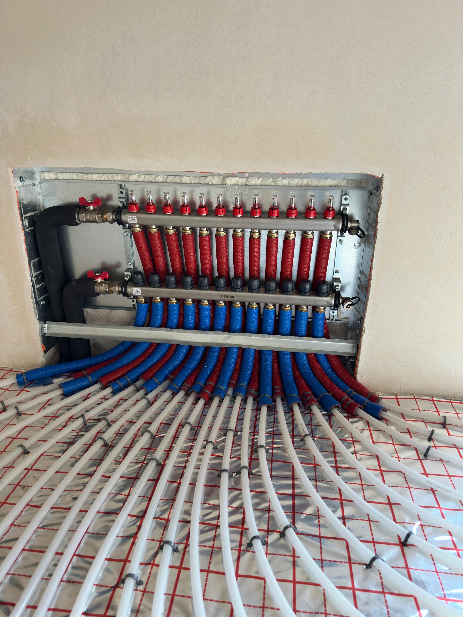 Rozdzielacz ogrzewania podłogowego z rurami PEX ułożonymi na folii aluminiowej z siatką, widoczne zawory regulacyjne i odpowietrzniki.