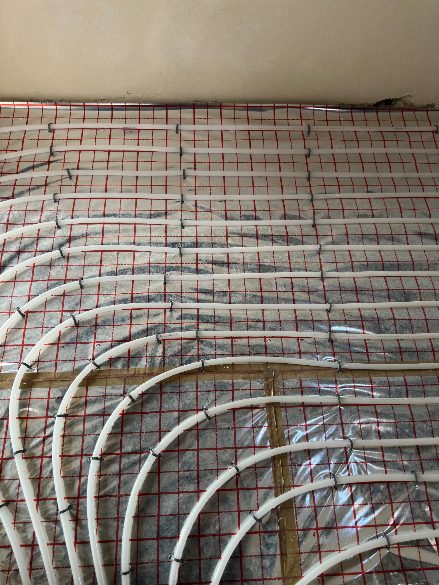 Białe rury instalacji grzewczej podłogowej, mocowane czarnymi klipsami do folii z czerwoną siatką, widok z góry na surową podłogę.