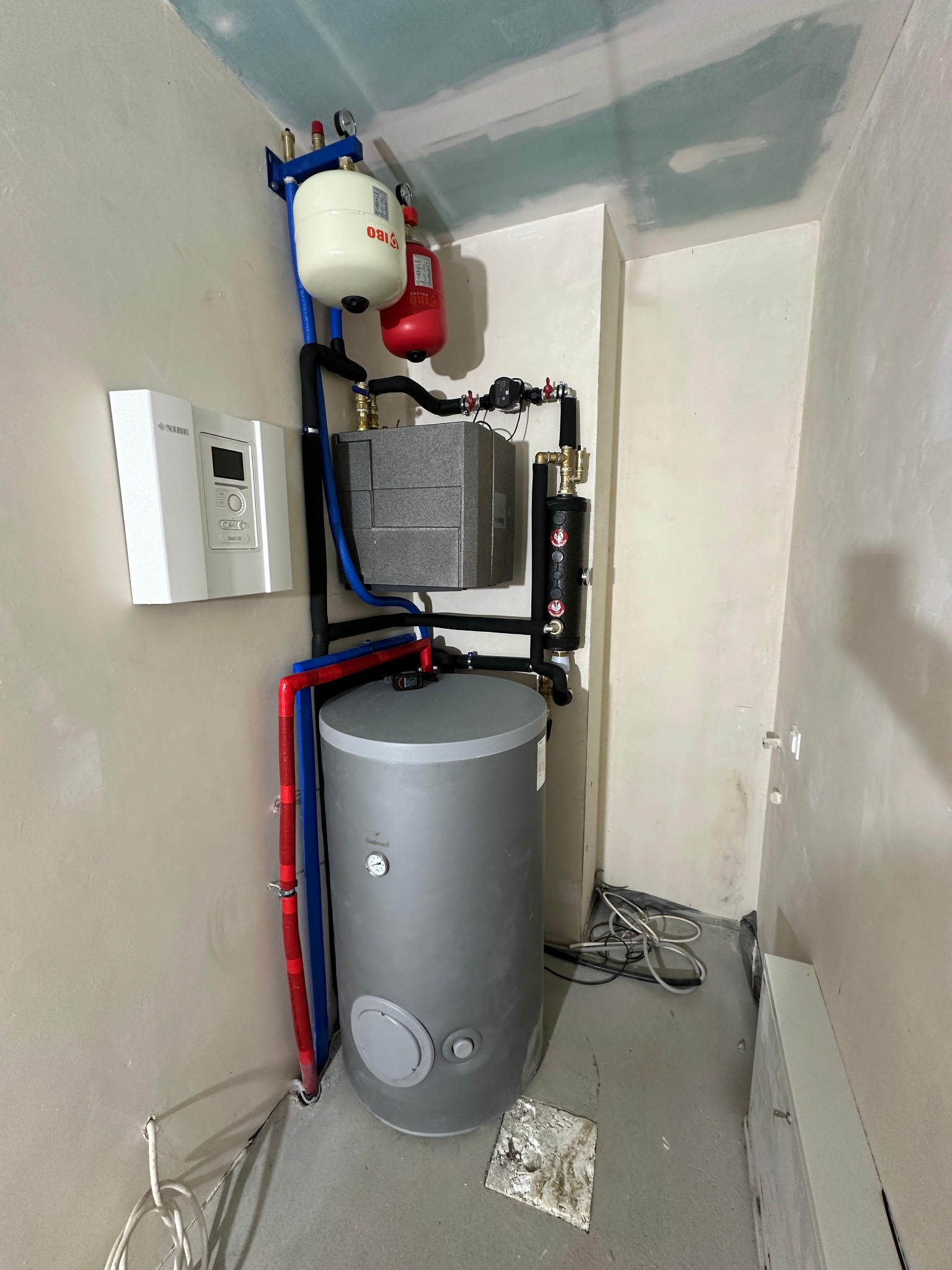 Szary zasobnik ciepłej wody użytkowej marki Galmet, podłączony do instalacji z czerwonymi i niebieskimi rurami, z białym panelem sterowania na ścianie, w pomieszczeniu w trakcie remontu.