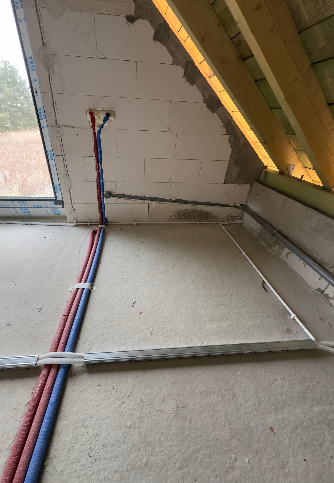 Wnętrze w trakcie prac instalacyjnych: czerwone i niebieskie rury prowadzone wzdłuż ściany z bloczków, mocowane do podłoża za pomocą uchwytów, widoczne również profile konstrukcyjne pod sufitem.