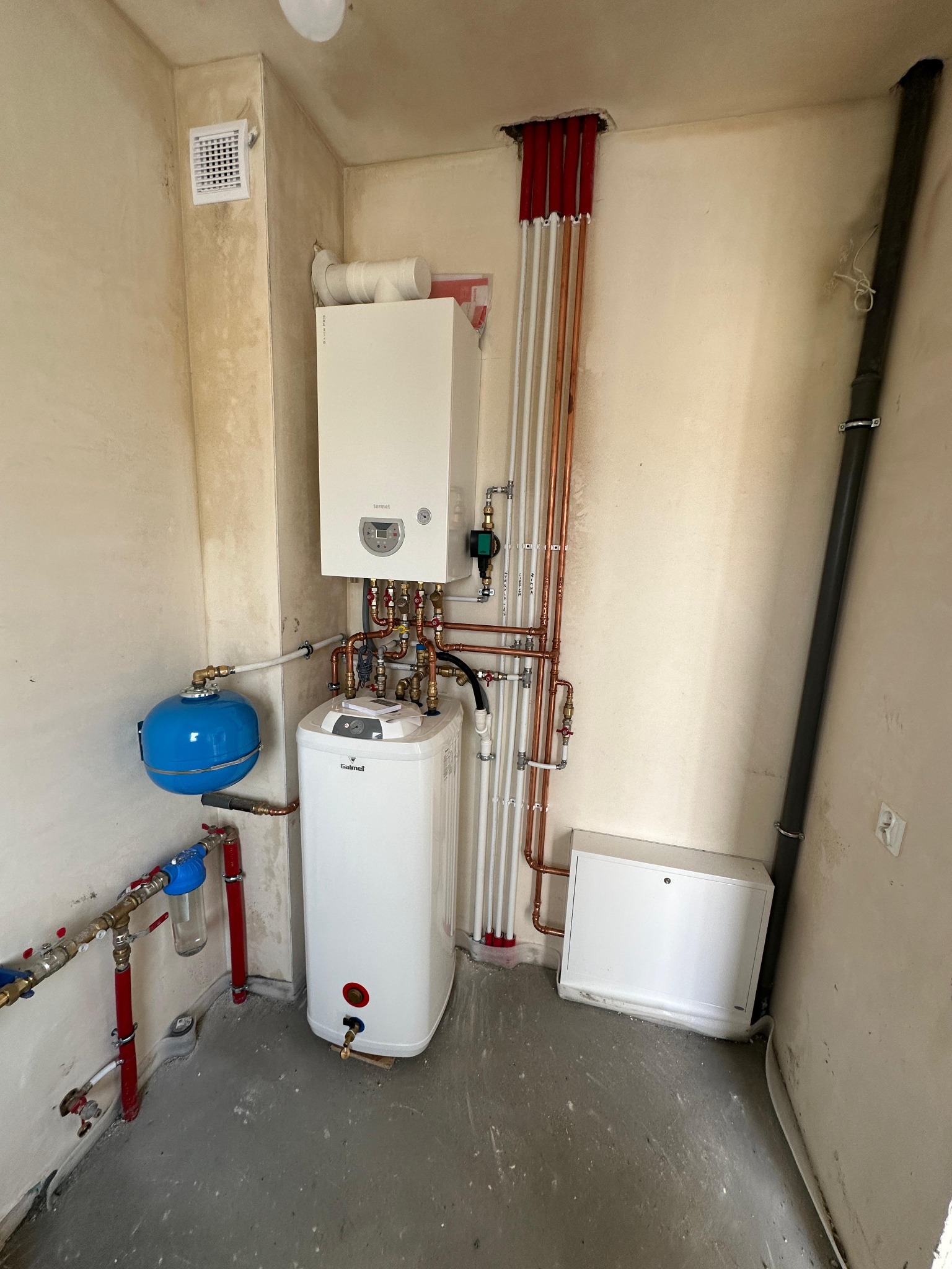 Zestaw urządzeń grzewczych w kotłowni: wiszący piec gazowy Termet, biały zasobnik Galmet, niebieskie naczynie przeponowe, rury miedziane w izolacji, szara rura kanalizacyjna przy ścianie.