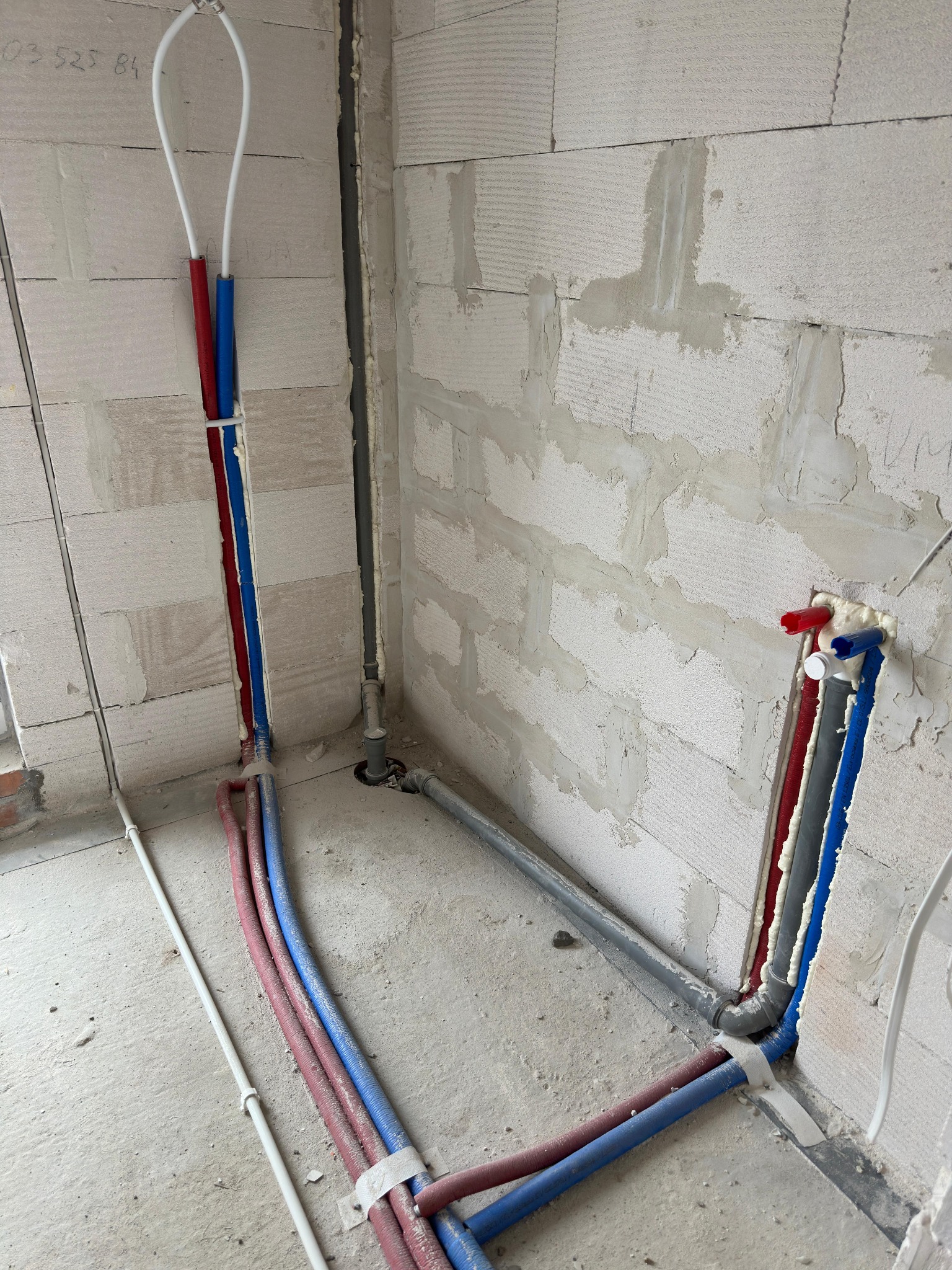 Instalacja wodna i kanalizacyjna w surowym wnętrzu, rury PEX czerwone i niebieskie prowadzone w ścianie z betonu komórkowego, widoczne pianowanie i mocowania.