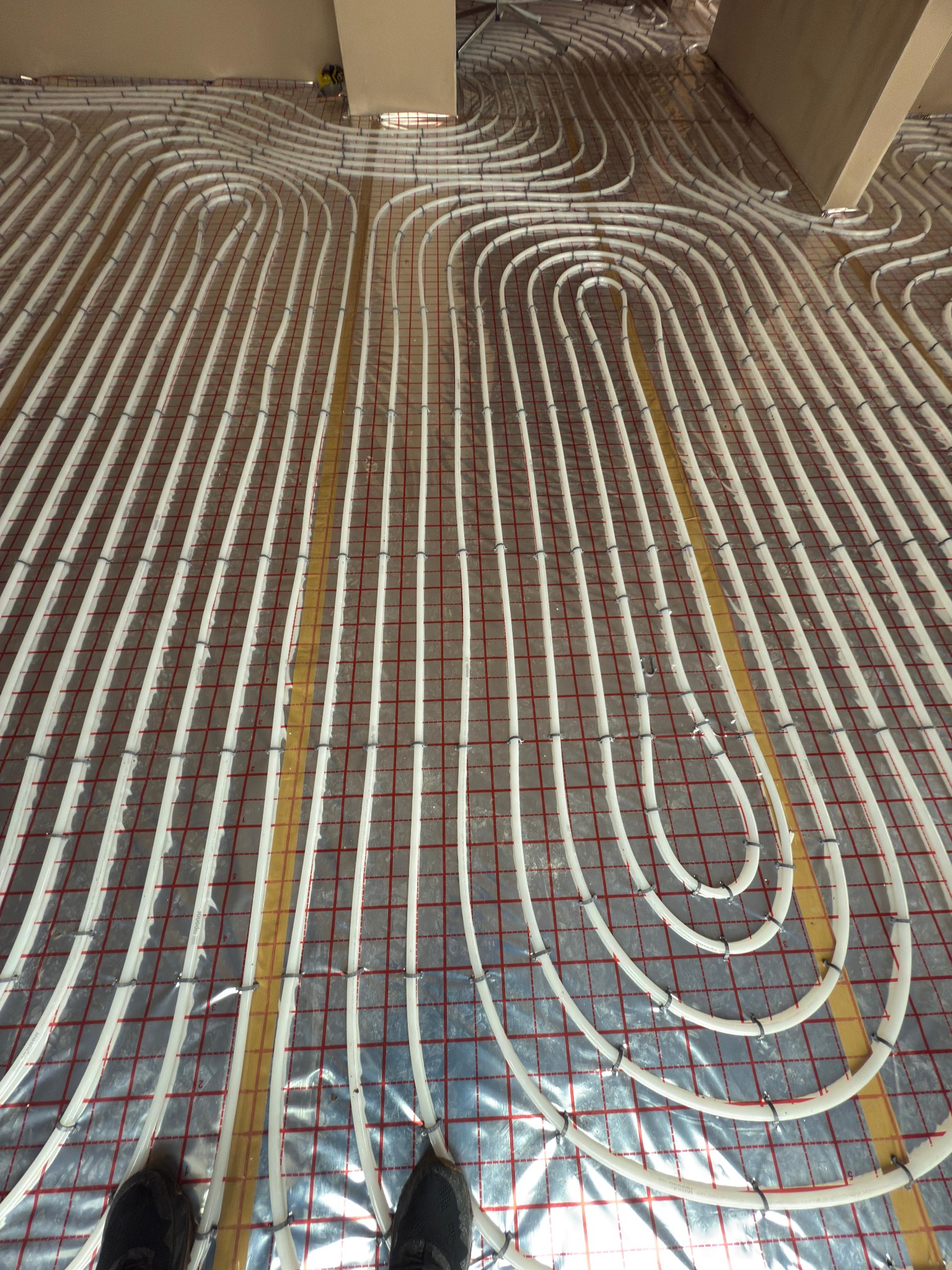 Białe rury systemu grzewczego podłogowego ułożone na izolacji z siatką w kolorze czerwonym, widoczne fragmenty butów instalatora w dolnej części kadru.