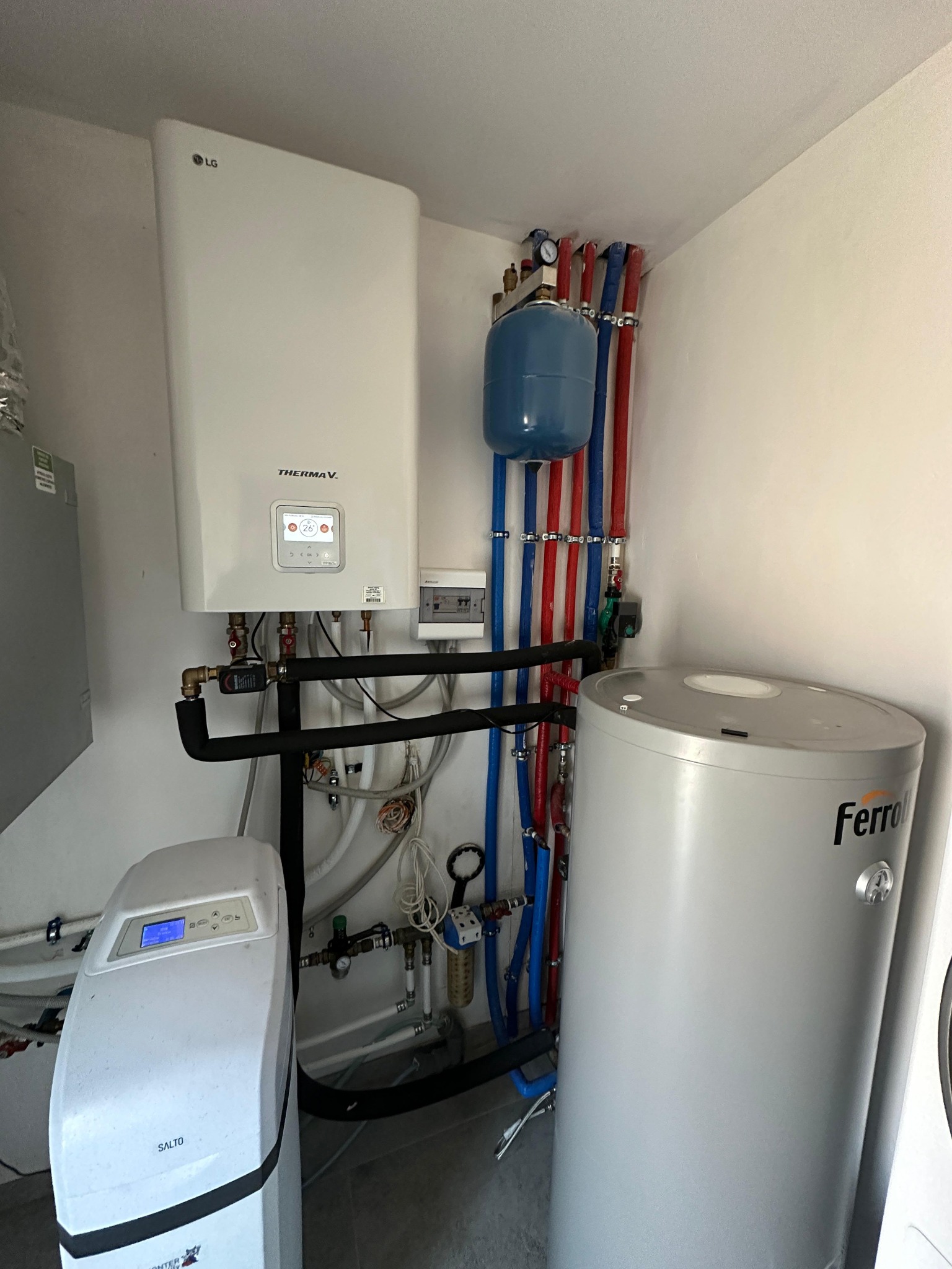 Biała jednostka wewnętrzna pompy ciepła LG Therma V, niebieski zbiornik wyrównawczy, szary zasobnik ciepłej wody Ferroli oraz zmiękczacz wody Salto w kotłowni z instalacją hydrauliczną z rur...
