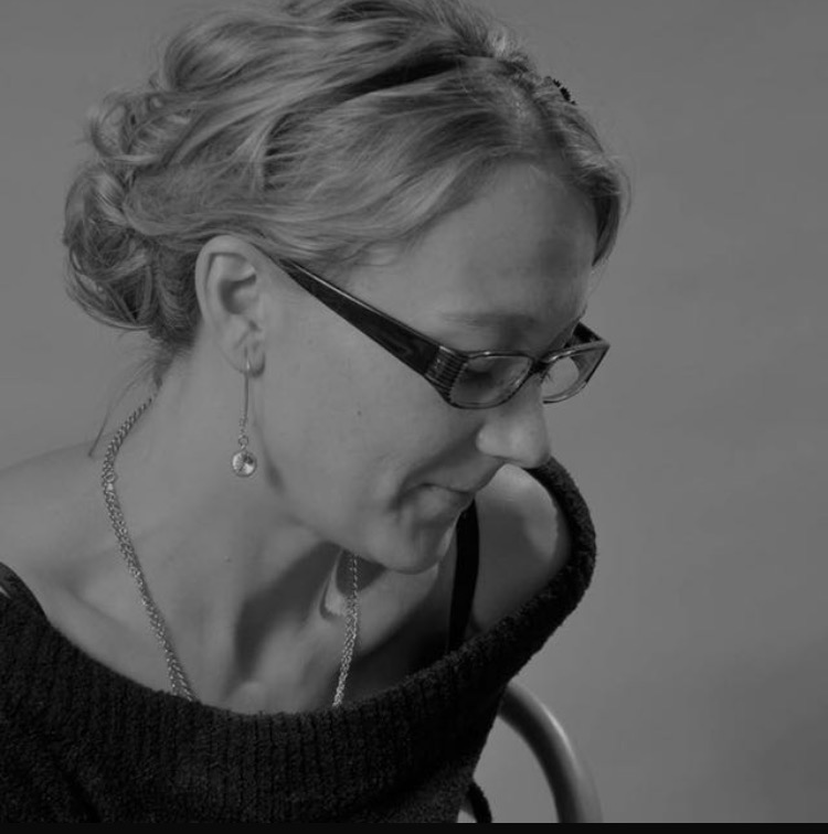 Czarno-białe zdjęcie kobiety w okularach, z upiętymi włosami i delikatną biżuterią, spoglądającej w dół. Ujęcie z profilu, z widocznym fragmentem krzesła.