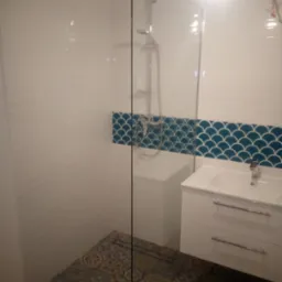 Nowoczesna łazienka z kabiną prysznicową typu walk-in, białą szafką pod umywalką i dekoracyjnymi płytkami na podłodze oraz niebieskimi płytkami rybie łuski nad szafką.