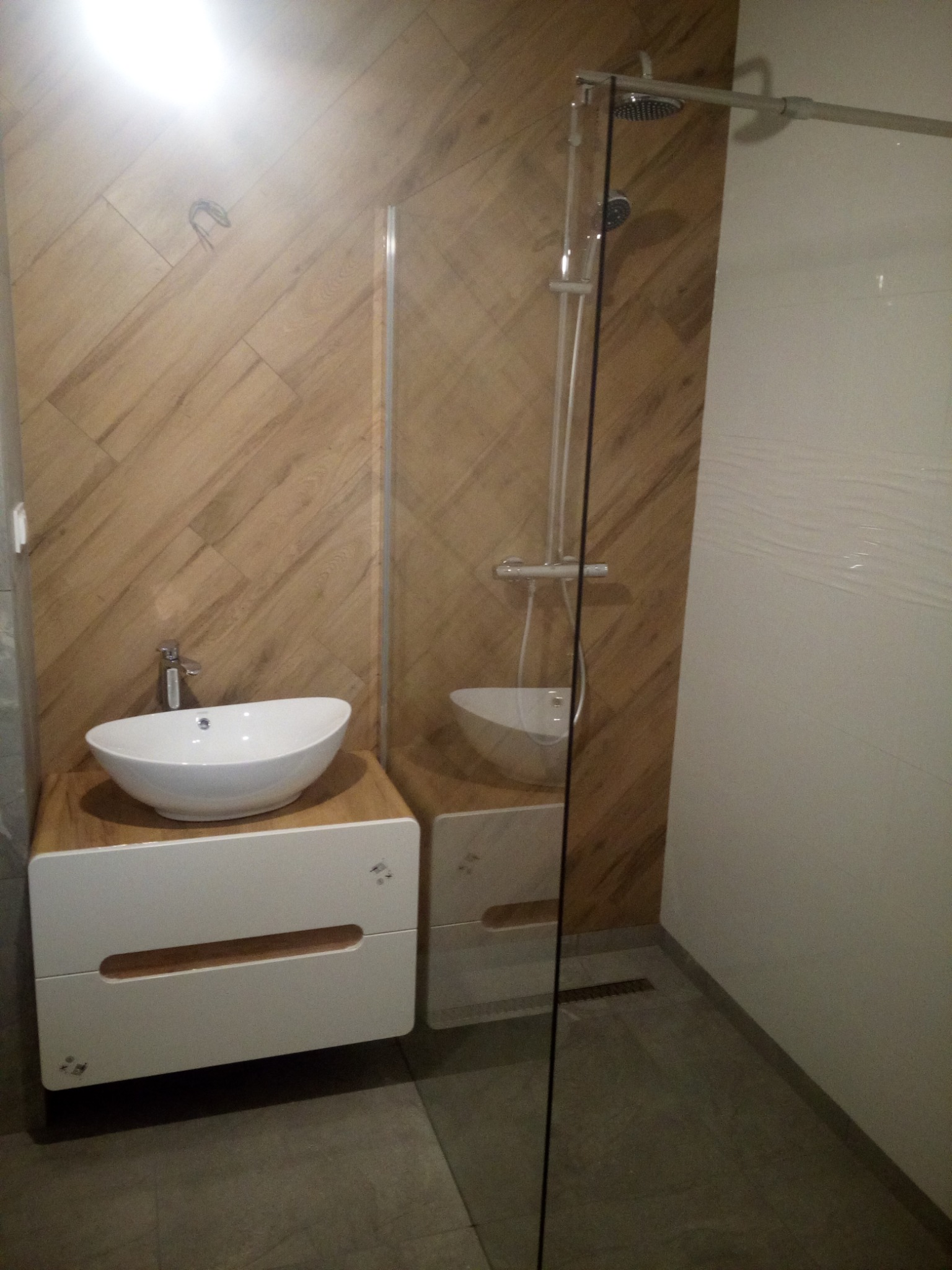 Nowoczesna łazienka z umywalką nablatową na szafce z drewnianym blatem, kabina prysznicowa ze szklaną ścianką i płytki imitujące drewno na ścianie.