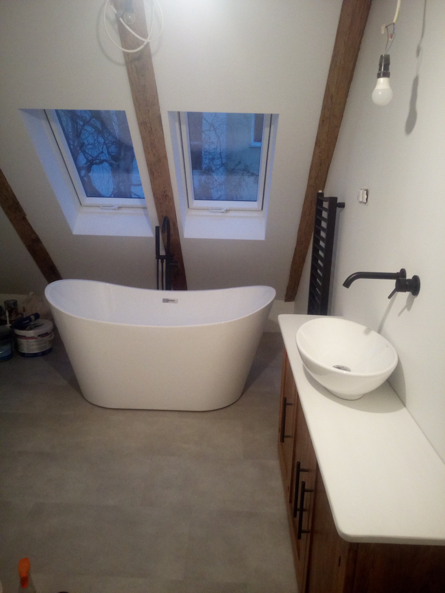 Biała wolnostojąca wanna i umywalka nablatowa w łazience na poddaszu z drewnianymi belkami stropowymi i oknami dachowymi, czarna armatura, grzejnik drabinkowy i surowa żarówka.