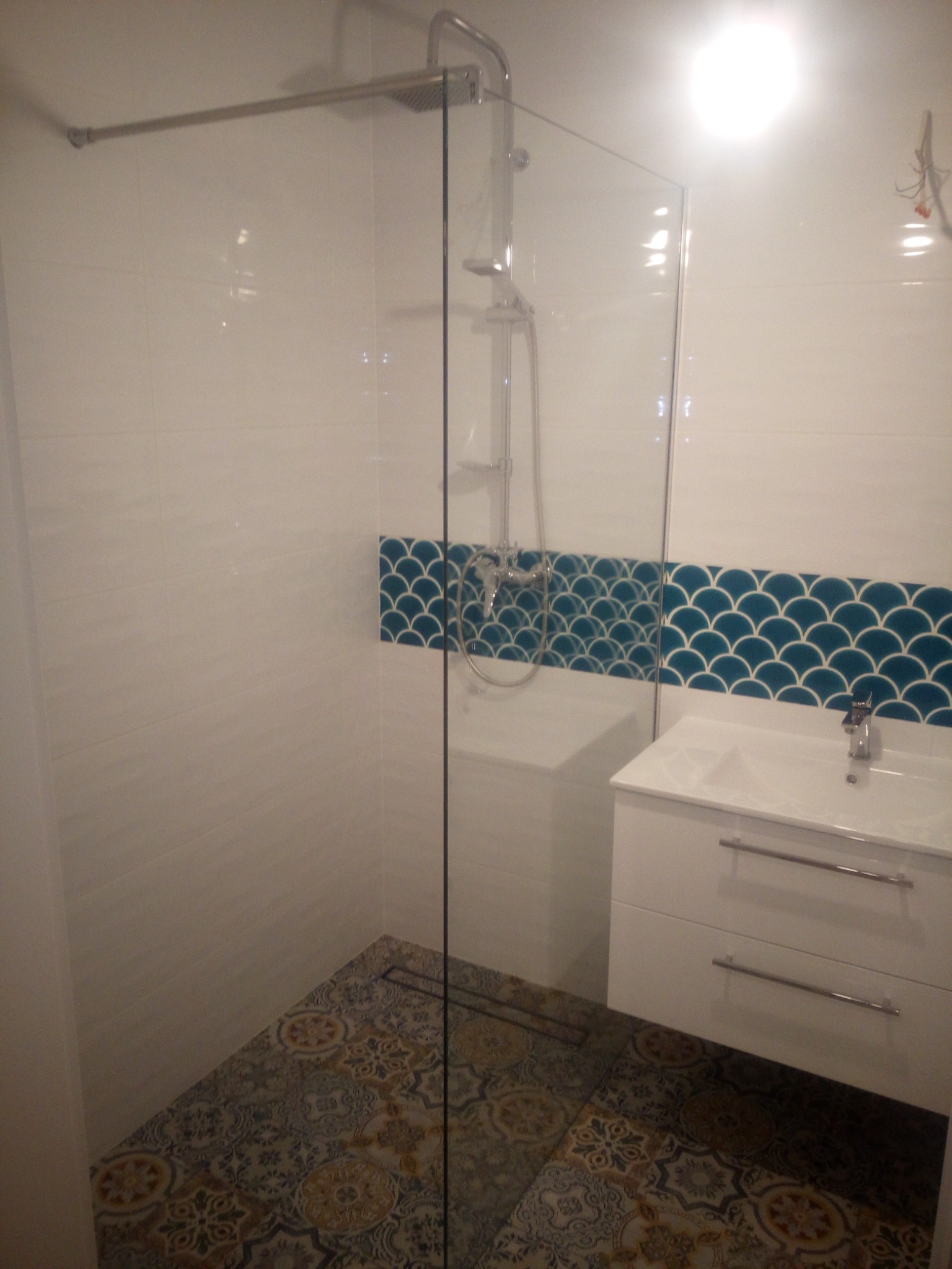 Nowoczesna łazienka z kabiną prysznicową typu walk-in, białą szafką pod umywalką i dekoracyjnymi płytkami na podłodze oraz niebieskimi płytkami rybie łuski nad szafką.