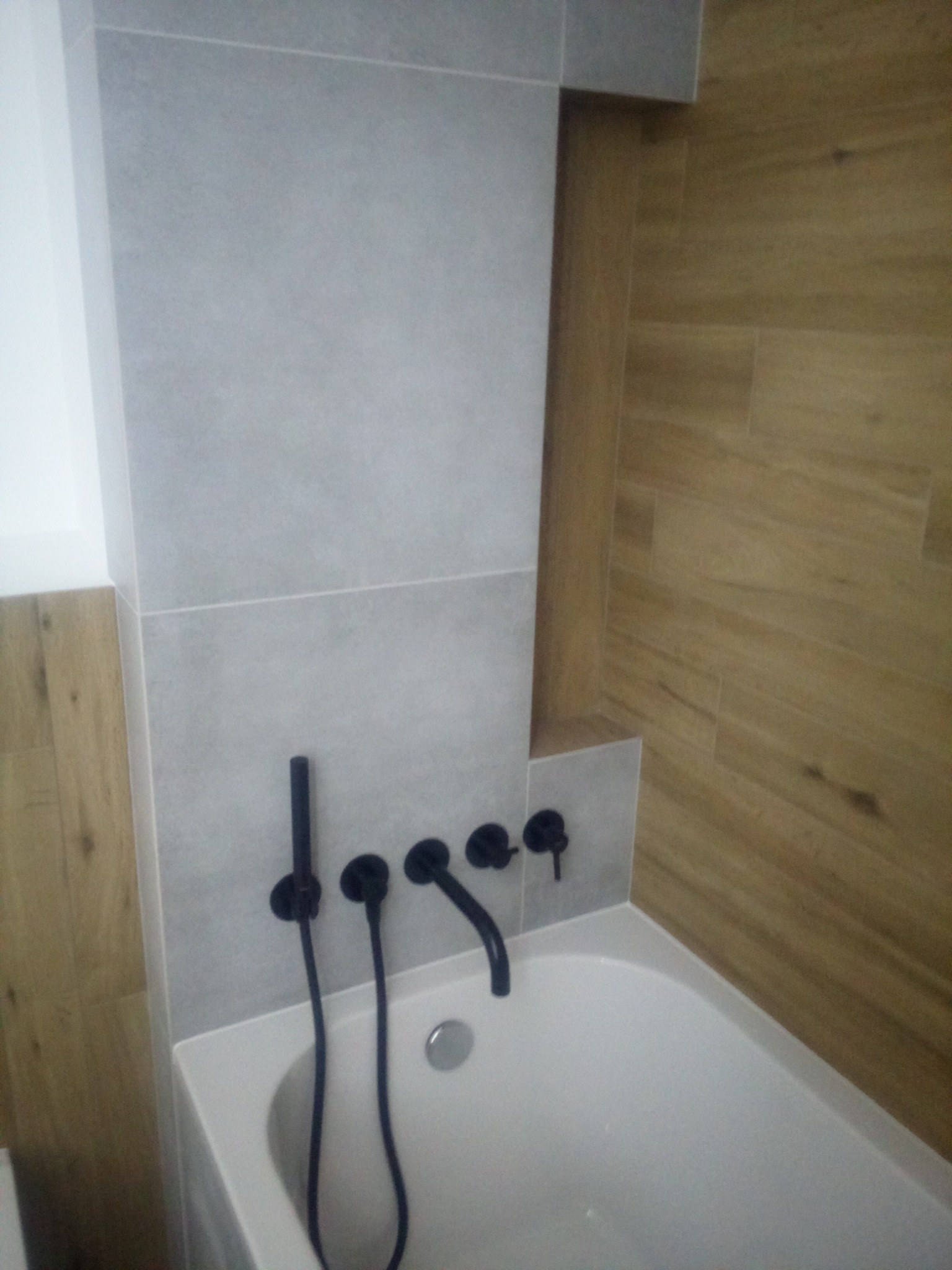 Wykończona łazienka z wanną, szarą płytką ścienną i drewnopodobną okładziną, armatura w kolorze czarnym.