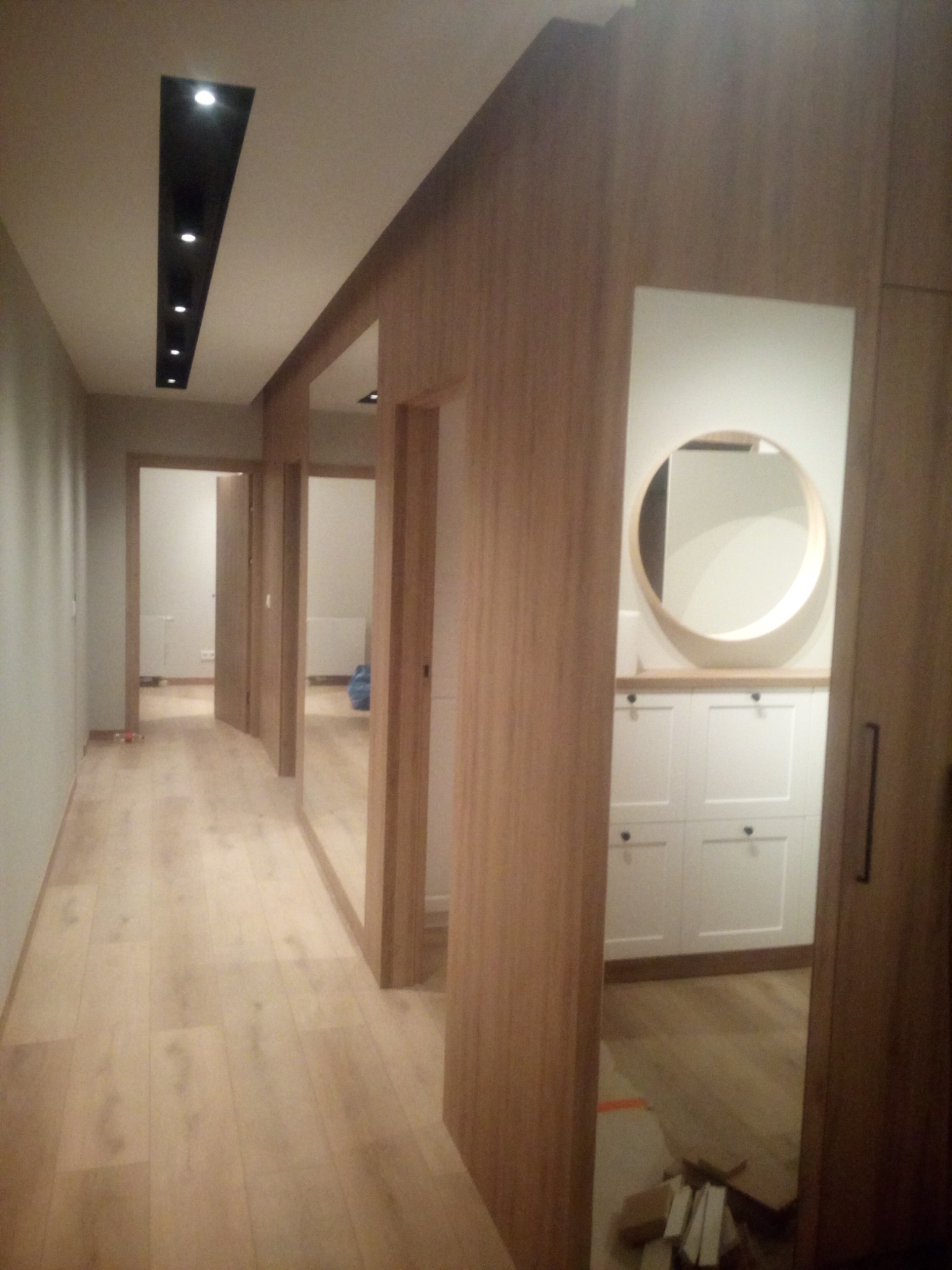 Korytarz z drewnianą podłogą i zabudową, widoczne drzwi wewnętrzne, okrągłe lustro nad białą komodą, czarny panel sufitowy z oświetleniem punktowym.