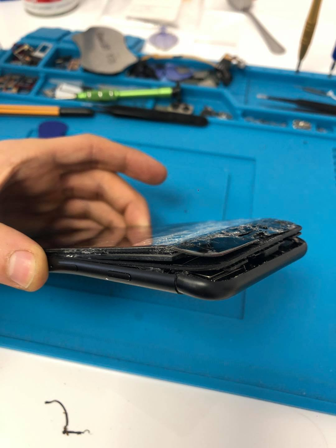 Uszkodzony, rozbity smartfon trzymany w dłoni nad niebieską matą serwisową z narzędziami w tle.