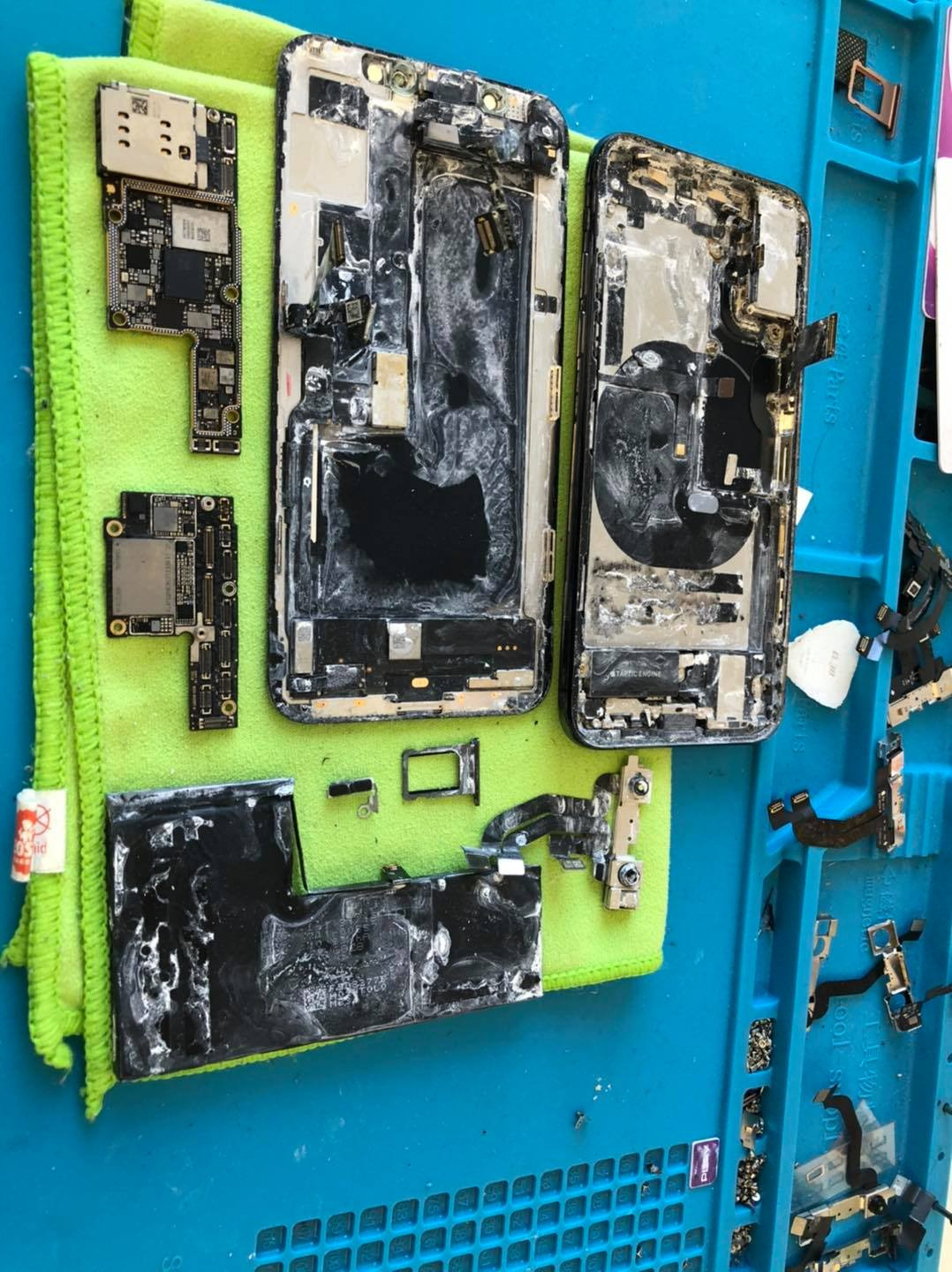 Rozłożone na części dwa uszkodzone smartfony iPhone, płyty główne, bateria i drobne elementy ułożone na zielonej ściereczce i niebieskiej macie serwisowej.