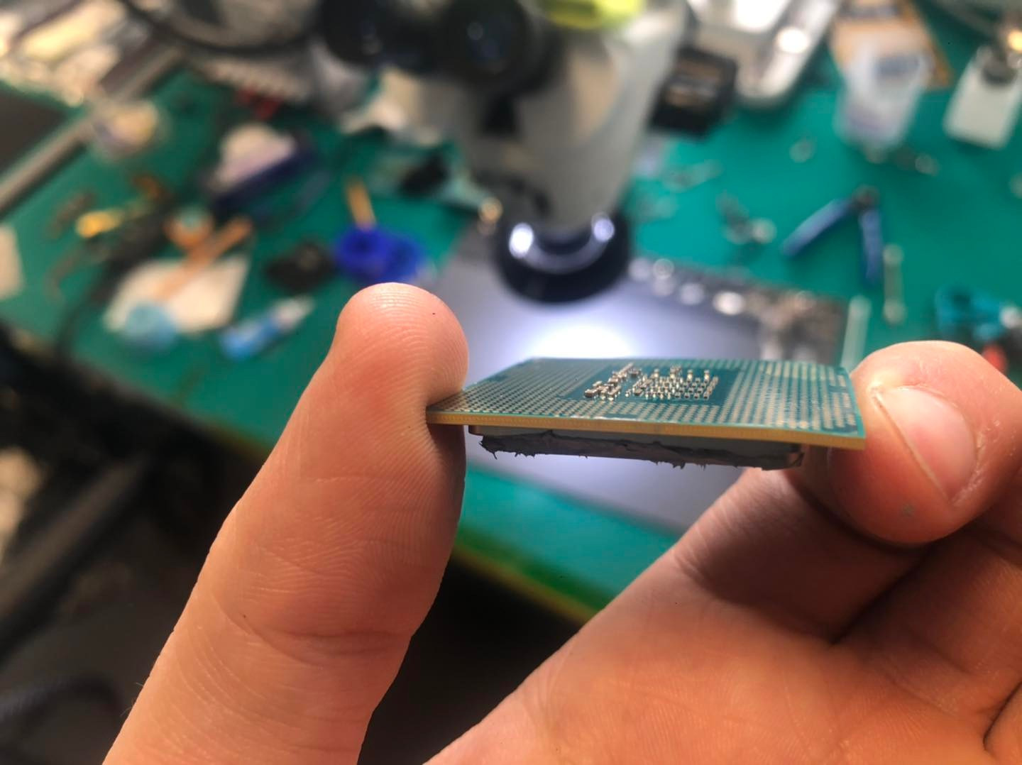 Palec trzyma procesor komputerowy w pobliżu mikroskopu, widoczny brud na krawędzi procesora i zielony blat roboczy z narzędziami w tle.