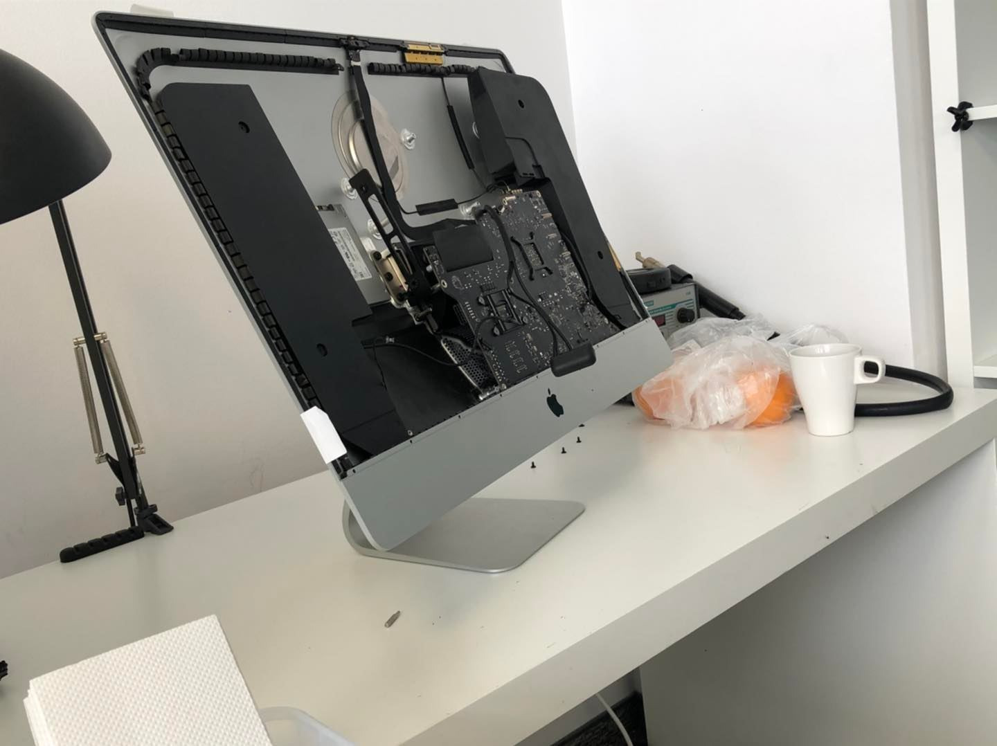 Otwarty iMac na białym biurku, widoczne wnętrze komputera, śrubki obok, lampka, kubek i pomarańcze w tle.