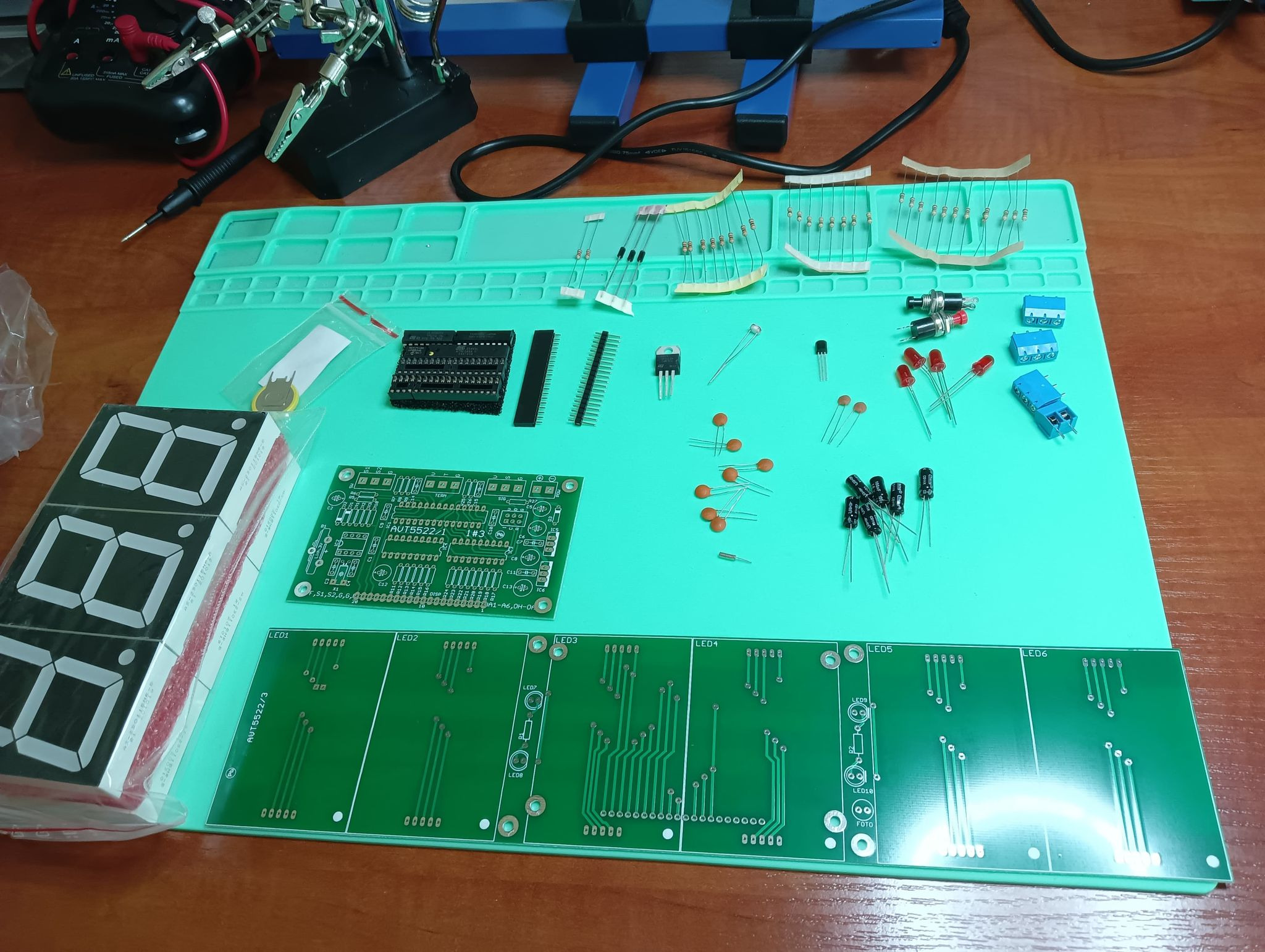 Elementy elektroniczne, w tym płytki drukowane, rezystory, kondensatory, diody LED i wyświetlacze siedmiosegmentowe, rozłożone na zielonej macie ESD, przygotowane do montażu.