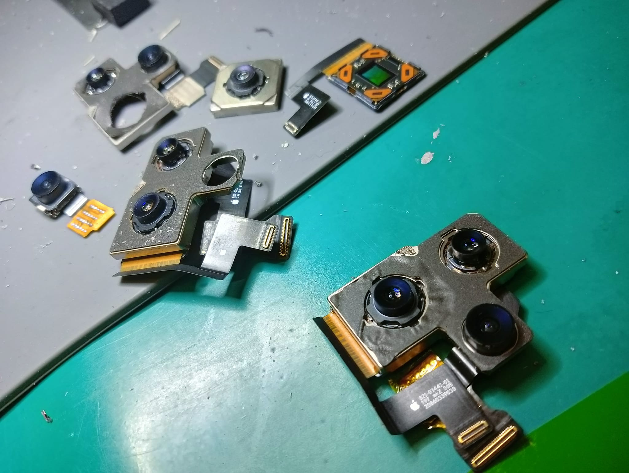 Rozmontowane moduły kamery smartfona, w tym potrójny moduł z widocznymi soczewkami i taśmami połączeniowymi, leżące na zielonej macie serwisowej.
