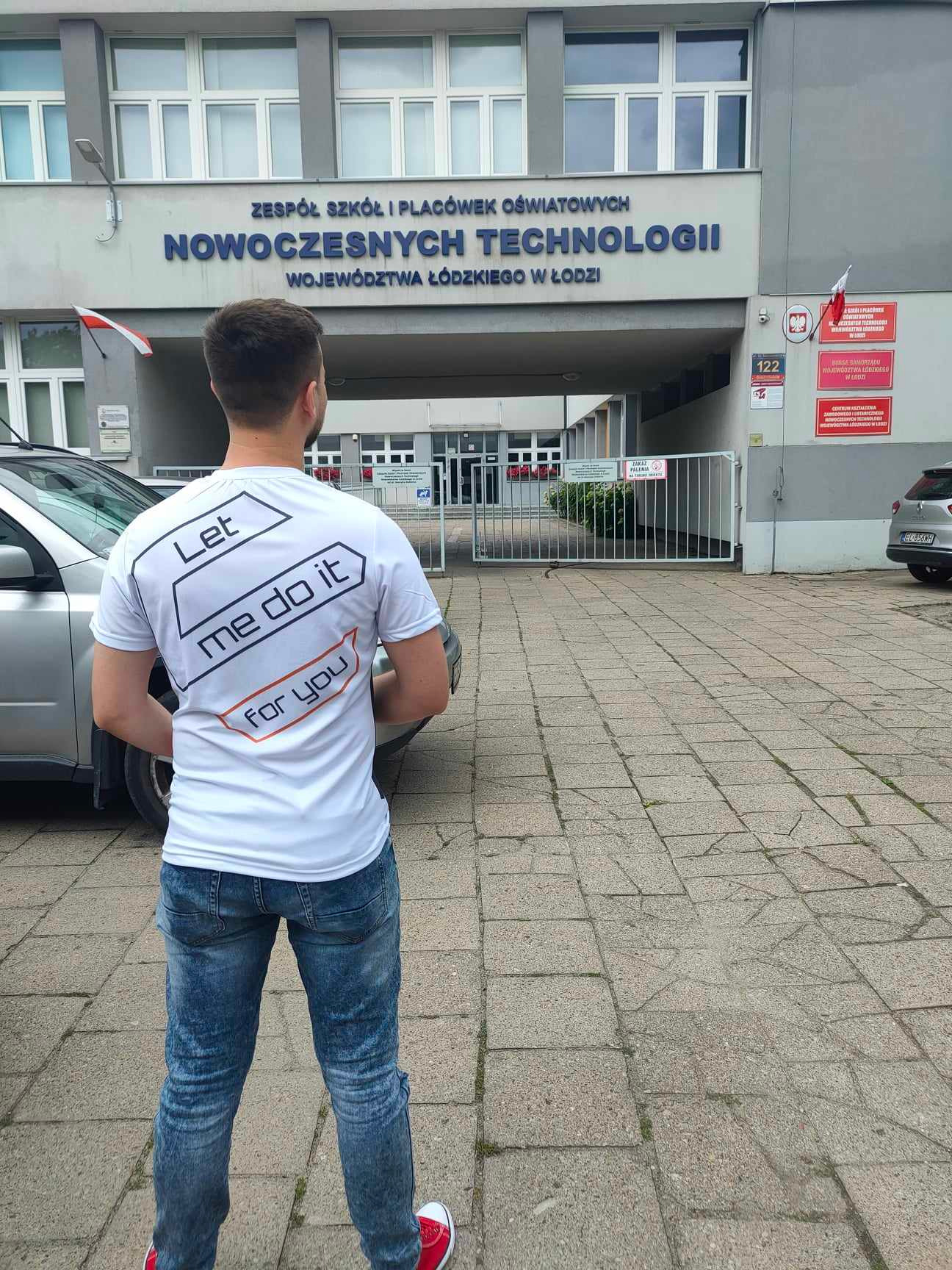 Mężczyzna w koszulce z napisem 'Let me do it for you' stojący tyłem przed budynkiem Zespołu Szkół i Placówek Oświatowych Nowoczesnych Technologii w Łodzi.