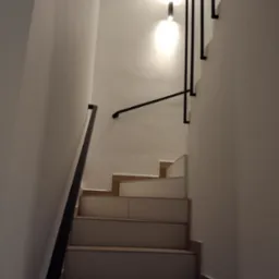 Klatka schodowa z białymi ścianami, drewnianymi stopniami i czarnymi, minimalistycznymi poręczami przymocowanymi do ściany oraz do sufitu za pomocą pionowych wsporników.