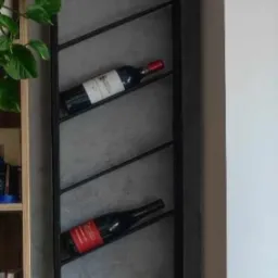 Metalowy regał na wino w stylu loft, przymocowany do szarej ściany, z czterema butelkami wina o różnych etykietach, obok półki z książkami i rośliny doniczkowej.