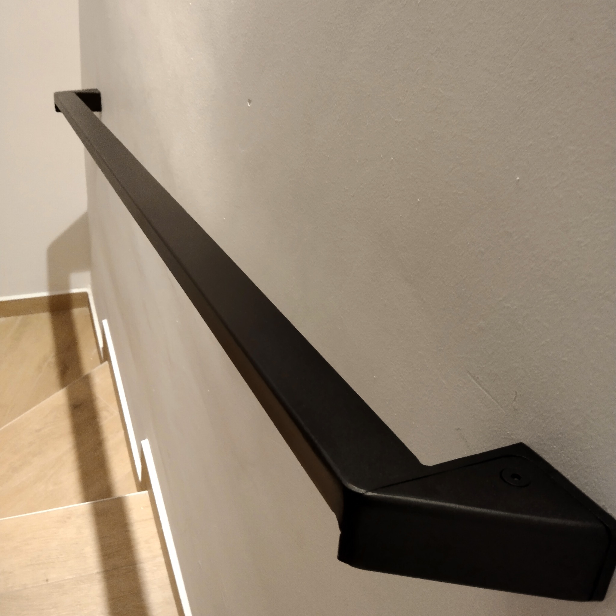 Czarna, metalowa poręcz zamontowana na ścianie wzdłuż schodów z jasnego drewna, z widocznymi śrubami mocującymi.