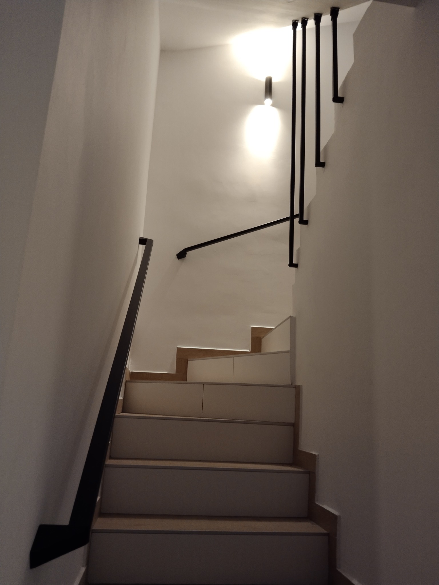 Klatka schodowa z białymi ścianami, drewnianymi stopniami i czarnymi, minimalistycznymi poręczami przymocowanymi do ściany oraz do sufitu za pomocą pionowych wsporników.